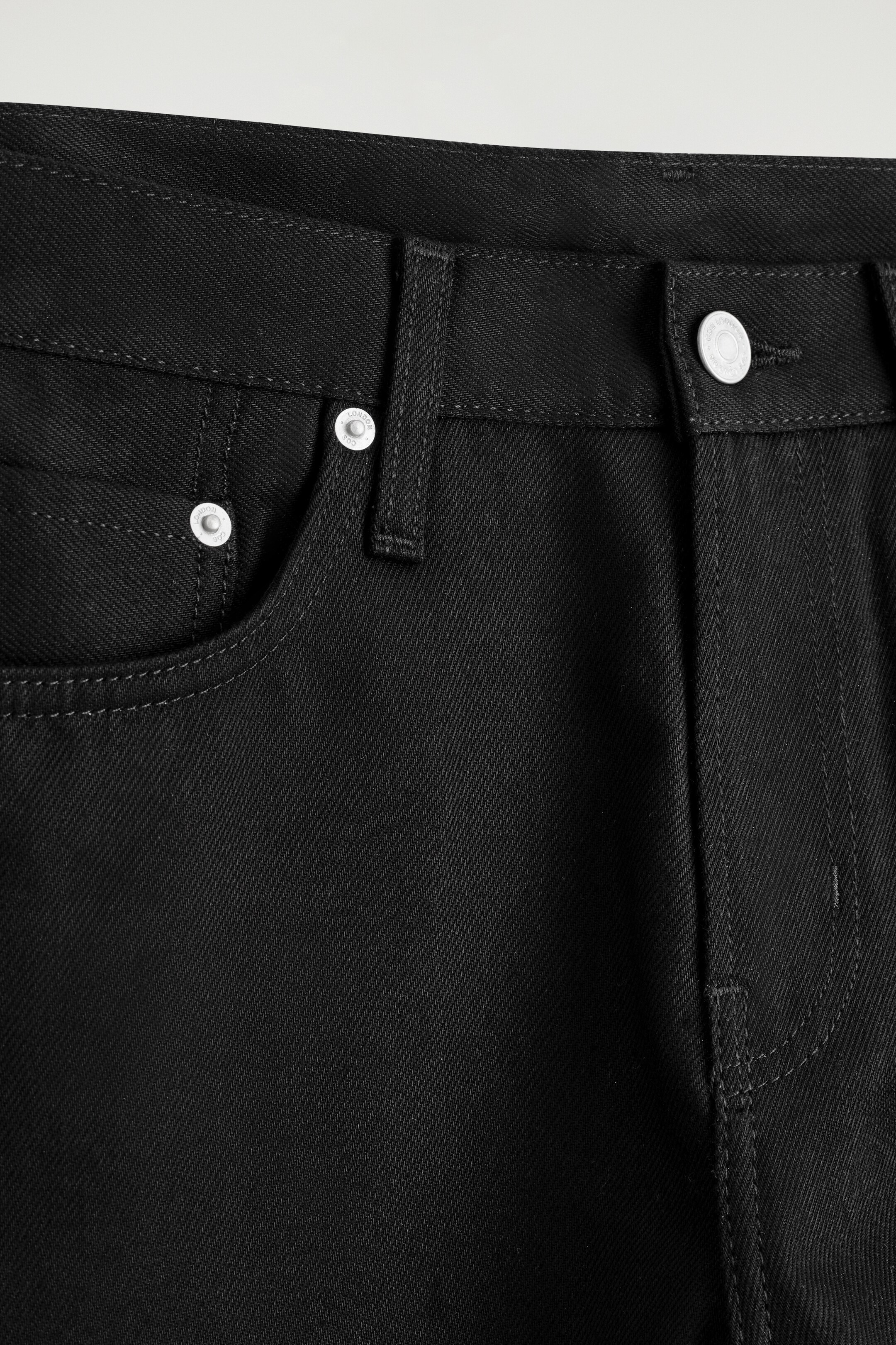 Grösseres Bild ansehen: Der obere Teil der schwarzen Jeans aus Denim wird angezeigt, mit einem Bund, Gürtelschlaufen und einer vorderen, abgerundeten Tasche mit einer Münztasche. Silberfarbene Nieten betonen die Taschen, und ein silberfarbener Knopf schließt den vorderen Hosenschlitz.