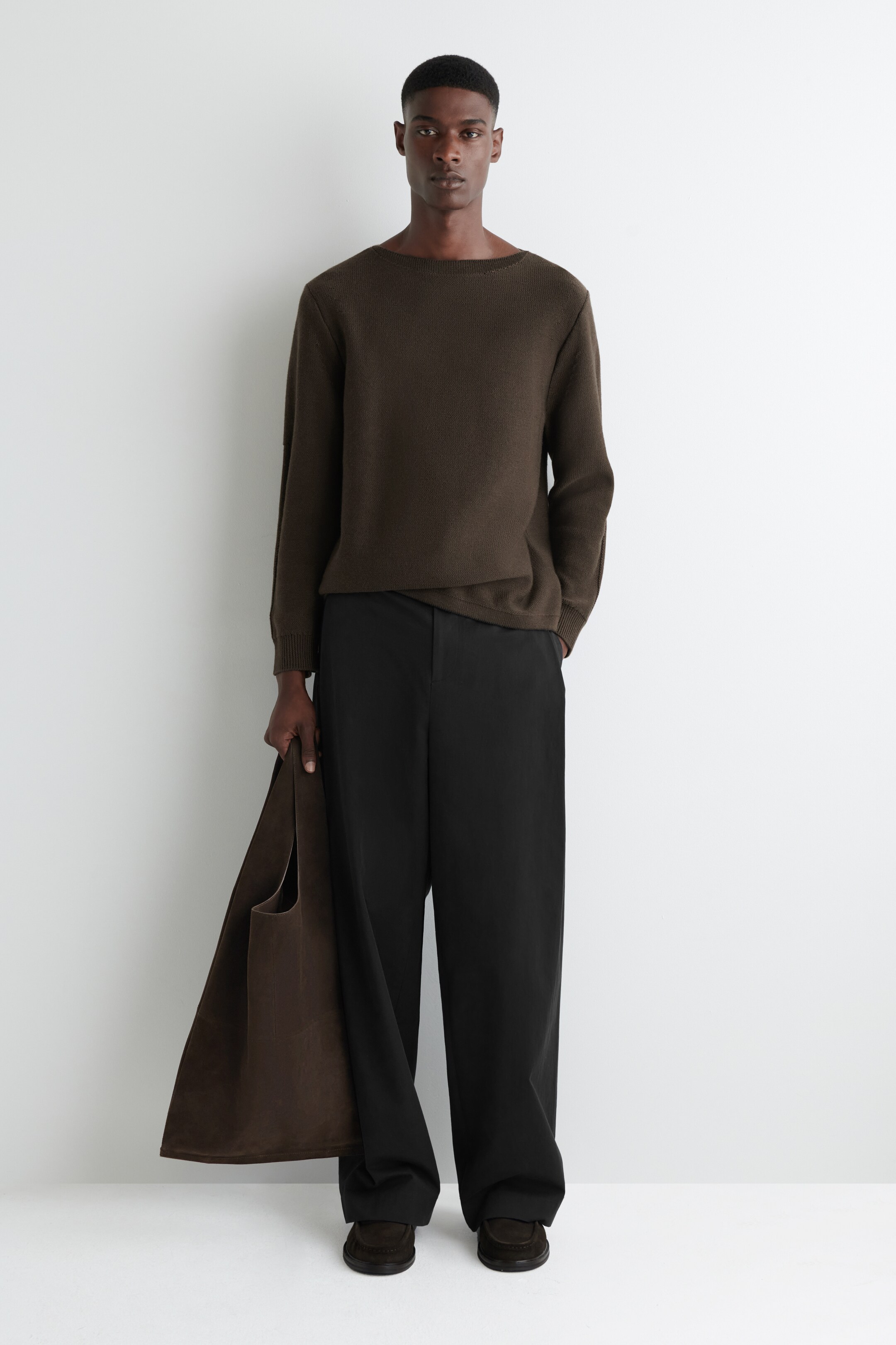 Agrandir l'image: Un pull en maille olive foncé avec un large décolleté est porté, assorti à un pantalon noir à jambes larges qui s'accumule sur des chaussures marron foncé sans lacets. Un grand sac fourre-tout marron foncé non structuré est tenu sur le côté.
