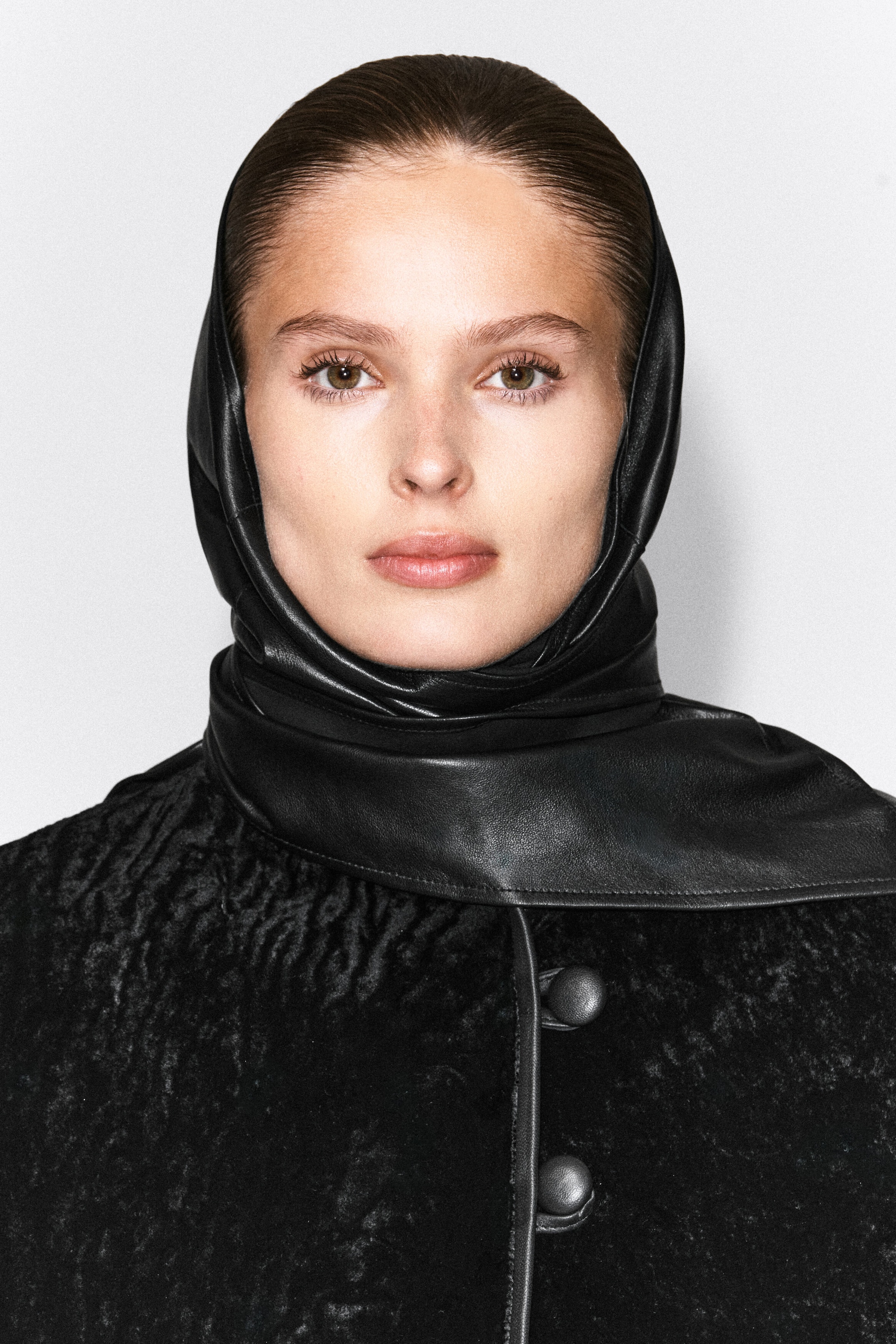 The model is wearing a black leather hood that fits closely around the head and wraps snugly around the neck. The hood is layered over a textured black coat with leather-trimmed button closures. The model is wearing a black leather hood that fits closely around the head and wraps snugly around the neck. The hood is layered over a textured black coat with leather-trimmed button closures.
