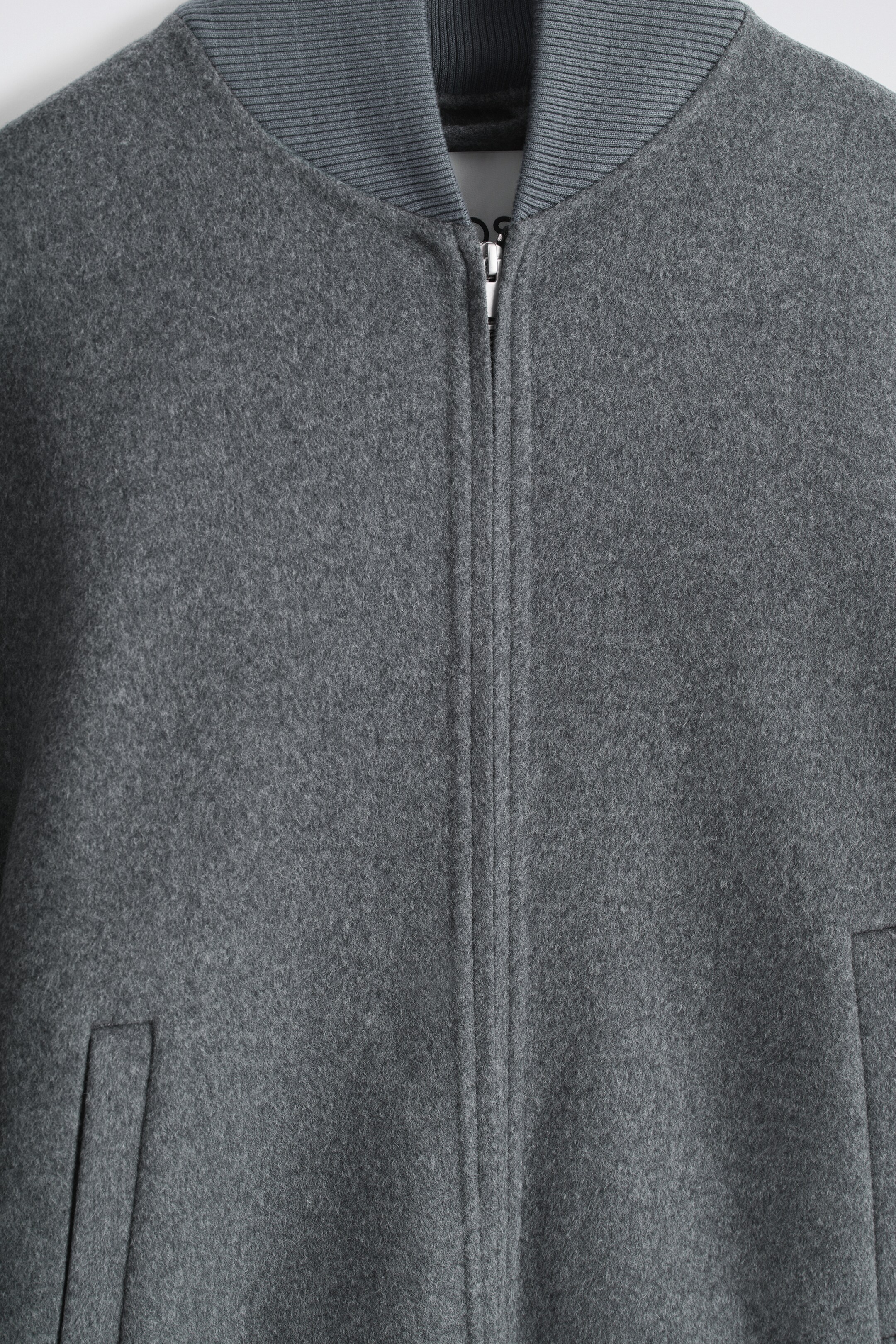View larger image: A grey bomber jacket is presenting its front, showcasing a soft, felted texture and a central silver-toned zip closure. It features a ribbed collar in a slightly darker grey and an angled welt pocket opening is visible on the lower right.