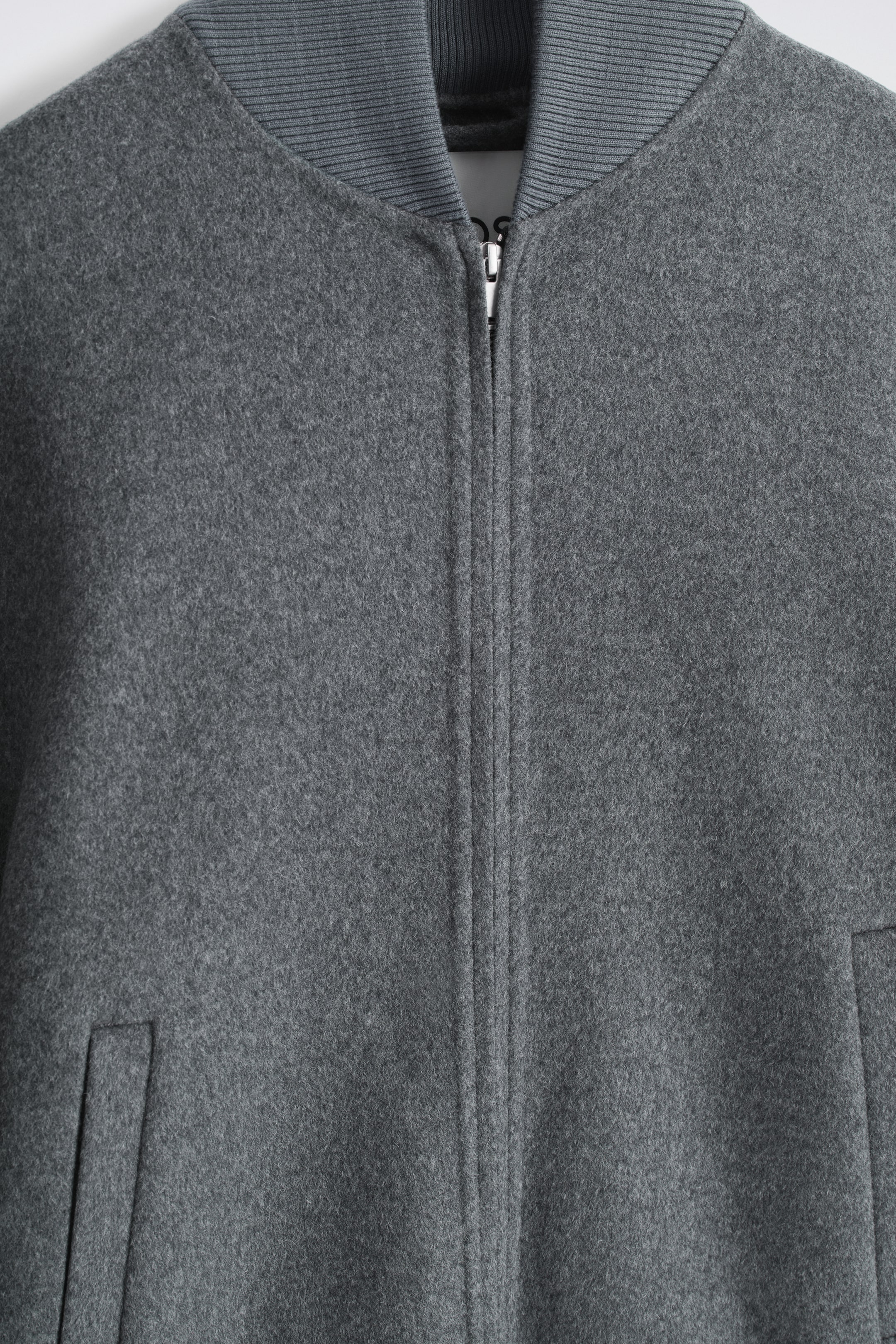 Ver imagen más grande: Una chaqueta bomber gris está presentando su parte frontal, mostrando una textura suave y afelpada y un cierre central con cremallera de tono plateado. Presenta un cuello acanalado en un gris ligeramente más oscuro y se puede ver una abertura de bolsillo ribeteada en ángulo en la parte inferior derecha.