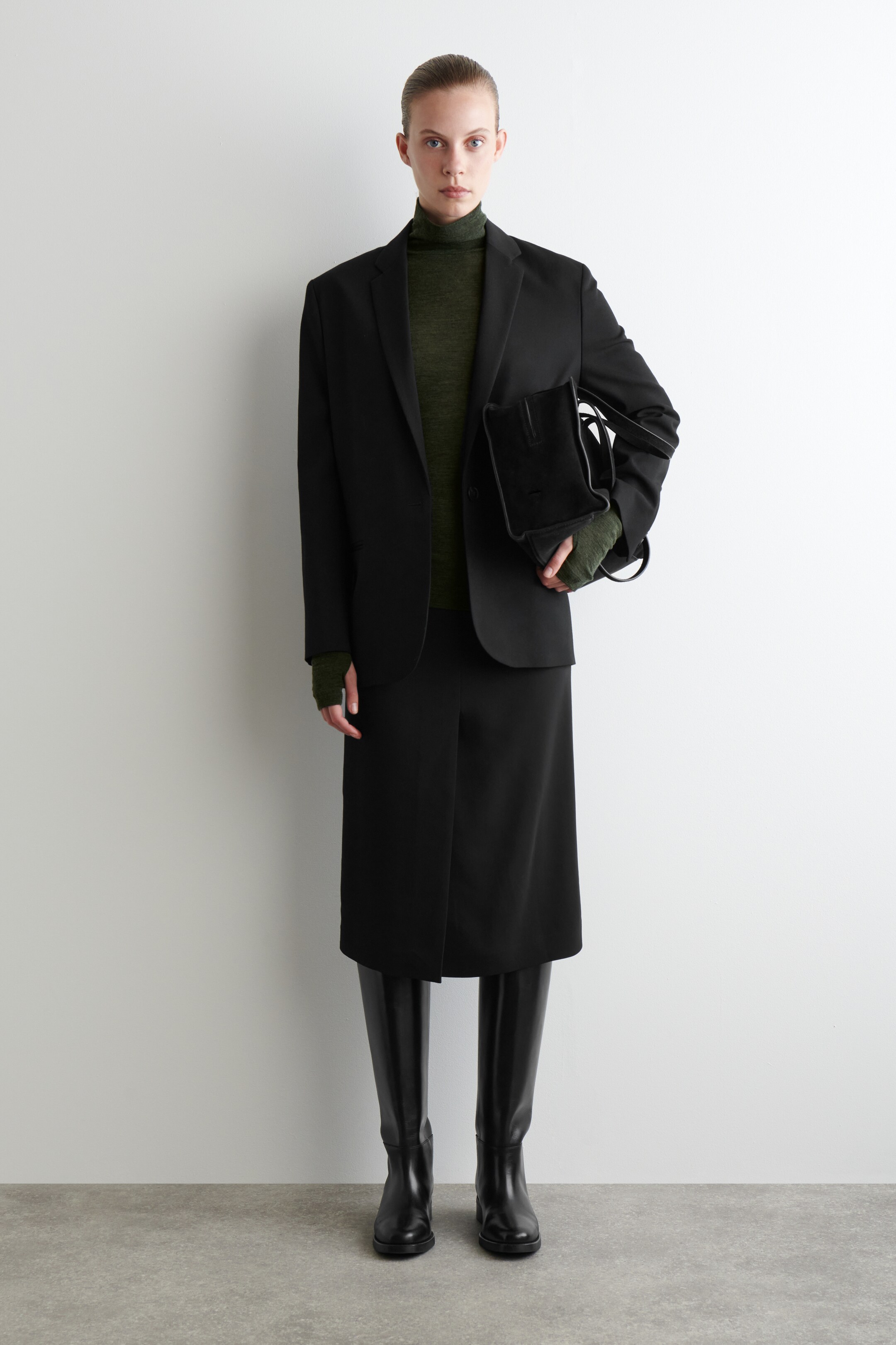 View larger image: A black single-breasted blazer with notch lapels is being worn front-facing, layered over an olive green fine-knit turtleneck. It is styled with a black midi-length skirt and black knee-high boots, while a black structured handbag is being held.