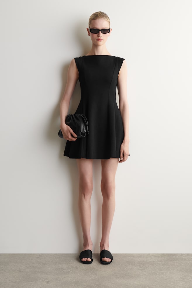 Une robe noire sans manches est présentée de face, avec un col bateau et un corsage ajusté qui s'évase en une jupe trapèze, se terminant au-dessus du genou. - 1