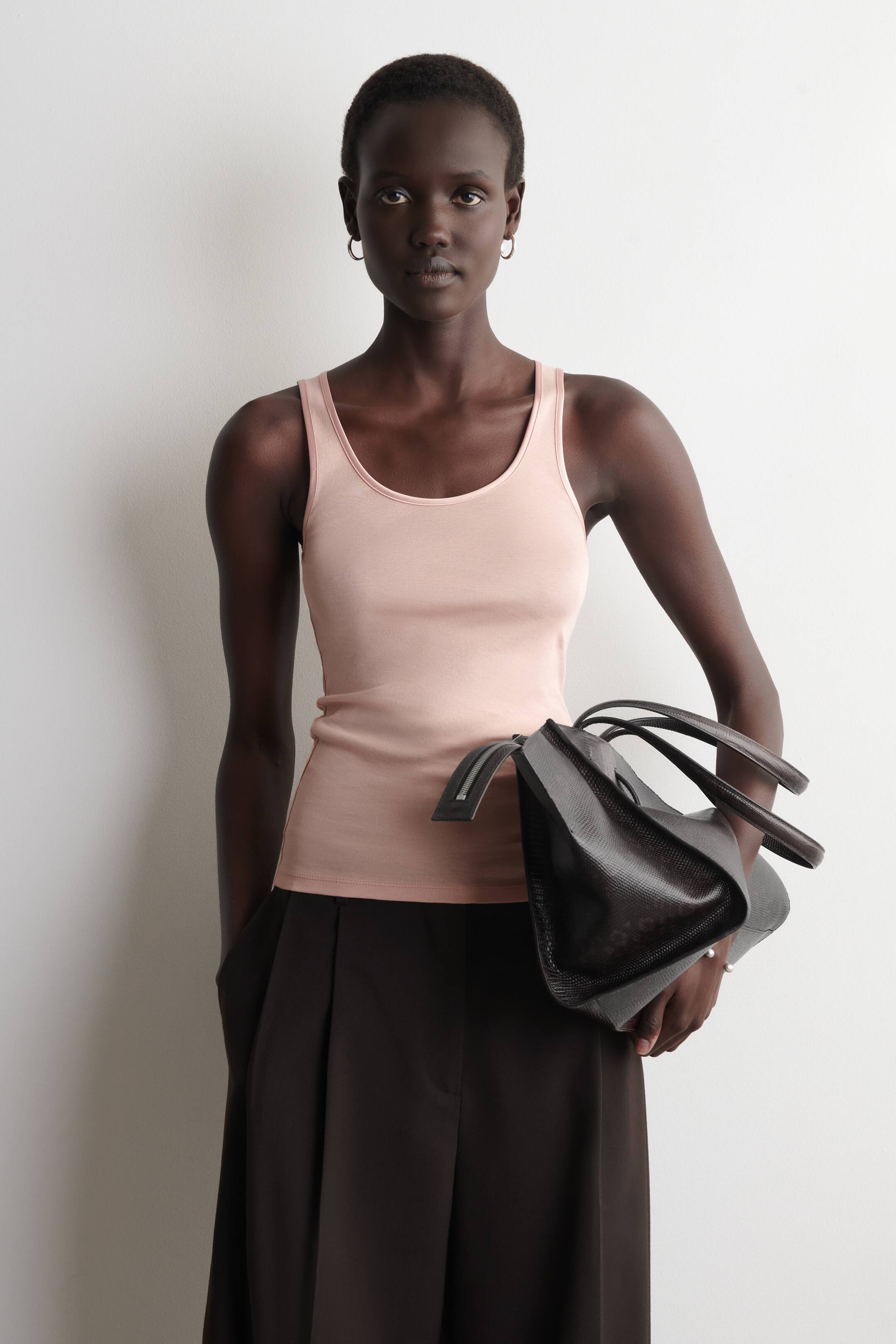 הצגת תמונה גדולה יותר: גופיית טנק בצבע ורוד בהיר עם מחשוף עגול נלבשת, מציגה סילואט מחוטב ומשולבת עם מכנסיים רחבים בצבע חום כהה עם קפלים קדמיים. תיק יד טקסטורלי בצבע חום כהה מוחזק, מציג ידית עליונה ופרט רוכסן גלוי.
