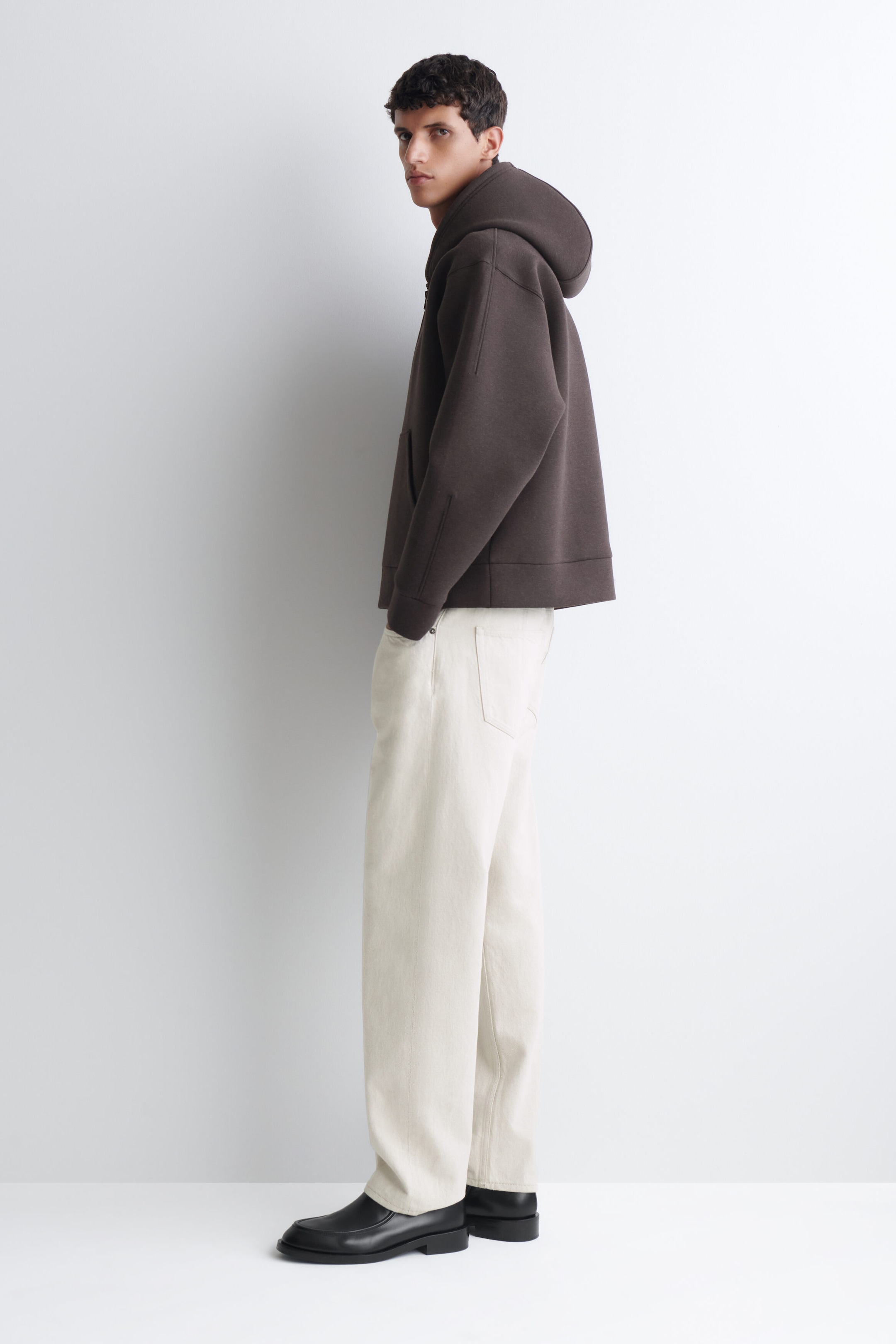 An oversized, dark brown scuba jersey zip-up hoodie with a structured hood and dropped shoulders is being worn by the model. The model is standing in profile, pairing the garment with cream-coloured trousers and black leather shoes. An oversized, dark brown scuba jersey zip-up hoodie with a structured hood and dropped shoulders is being worn by the model. The model is standing in profile, pairing the garment with cream-coloured trousers and black leather shoes.