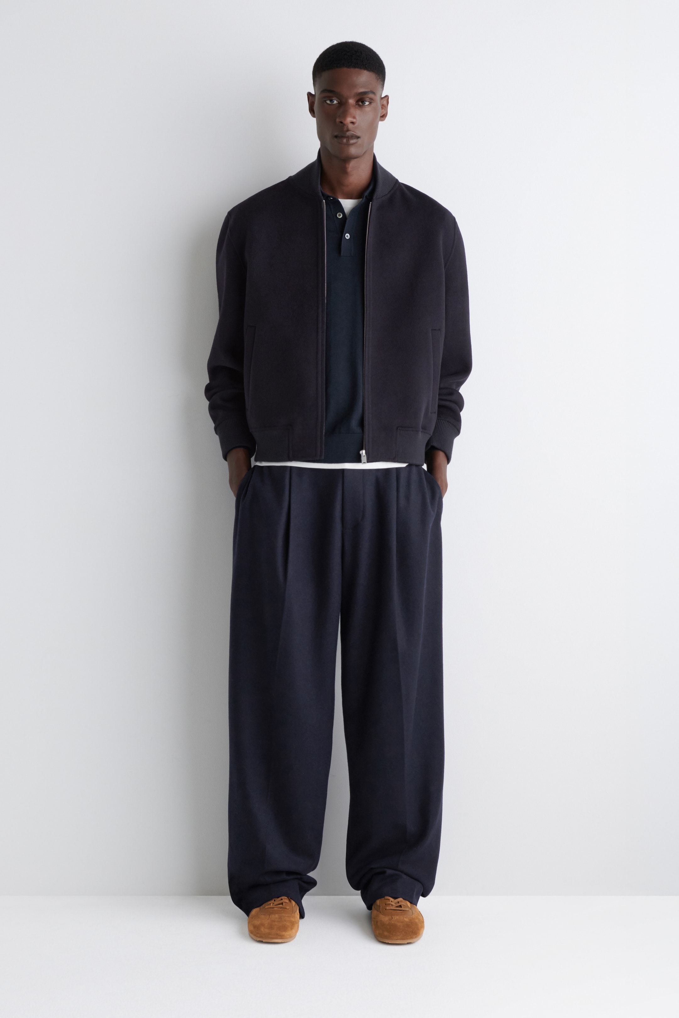 View larger image: An outfit featuring a dark navy bomber jacket, a dark navy polo shirt, and wide-leg dark navy trousers is being presented from a front view. The bomber jacket is showcasing a full-zip closure with a ribbed collar, cuffs, and hem. The trousers are exhibiting a pleated front and a relaxed fit, while a white t-shirt is visible layered at the hem, and tan suede shoes are completing the ensemble.