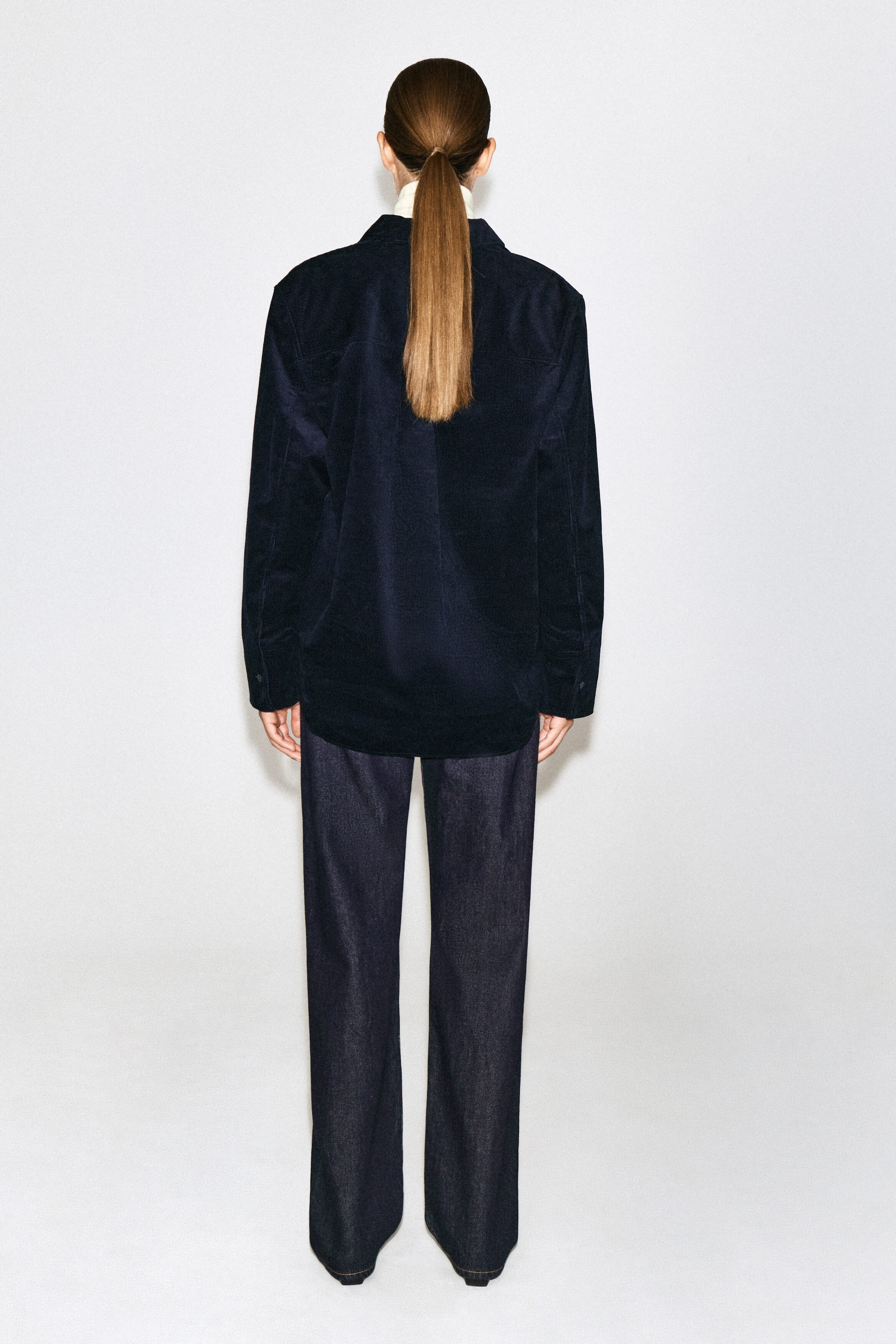 The model is wearing a dark navy cotton corduroy shirt from the back, highlighting its relaxed fit, long sleeves, and back yoke. The shirt is styled over a light-colored high-neck top and paired with dark denim trousers. The model is wearing a dark navy cotton corduroy shirt from the back, highlighting its relaxed fit, long sleeves, and back yoke. The shirt is styled over a light-colored high-neck top and paired with dark denim trousers.