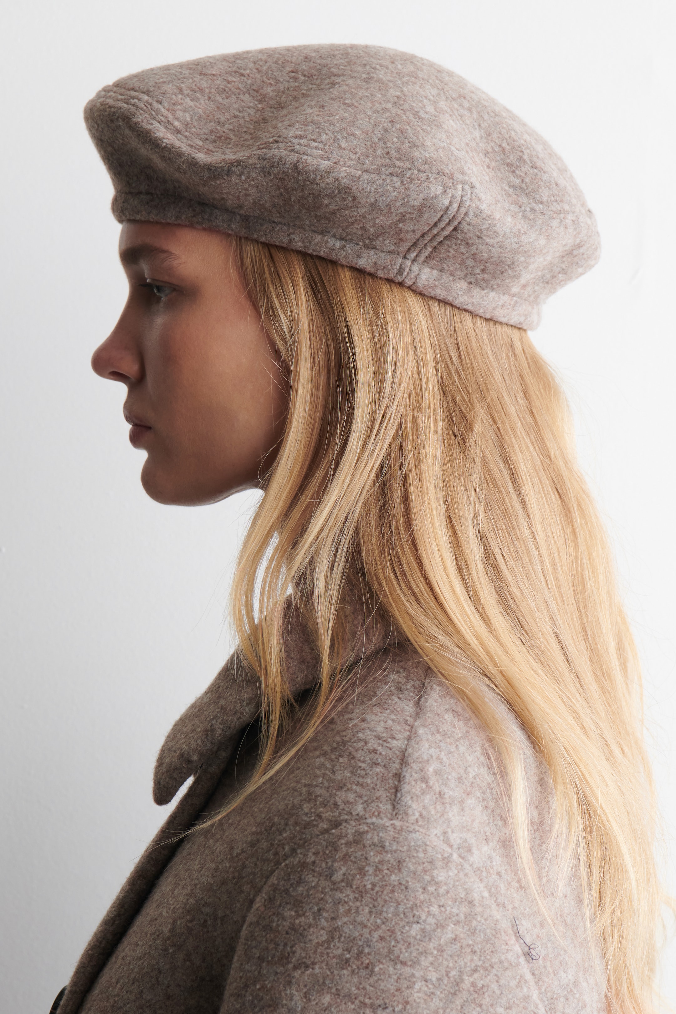 Agrandir l'image: Un béret classique de couleur mélange-beige est porté, incliné vers la gauche. Il présente une couronne douce et arrondie avec une finition subtile et texturée, ainsi qu'une bande plate distincte autour de la base.
