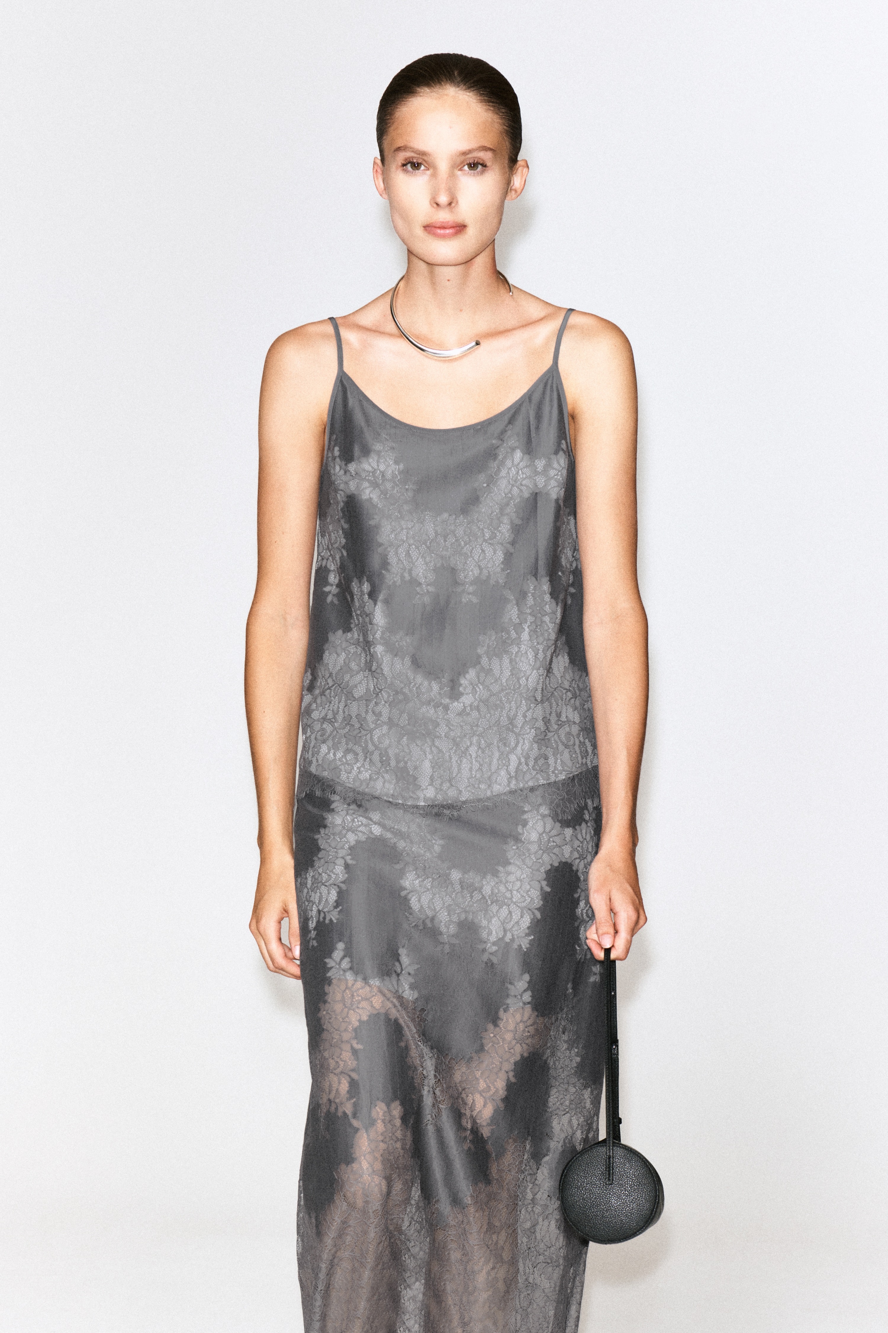 Un top cami grigio con inserto in pizzo Ã¨ mostrato frontalmente, con sottili spalline spaghetti e scollo tondo. Il top presenta un tessuto semi-trasparente con raffinati sovrapposizioni in pizzo floreale che si estendono su tutto il corpo e terminano in un orlo smerlato in pizzo eyelash.