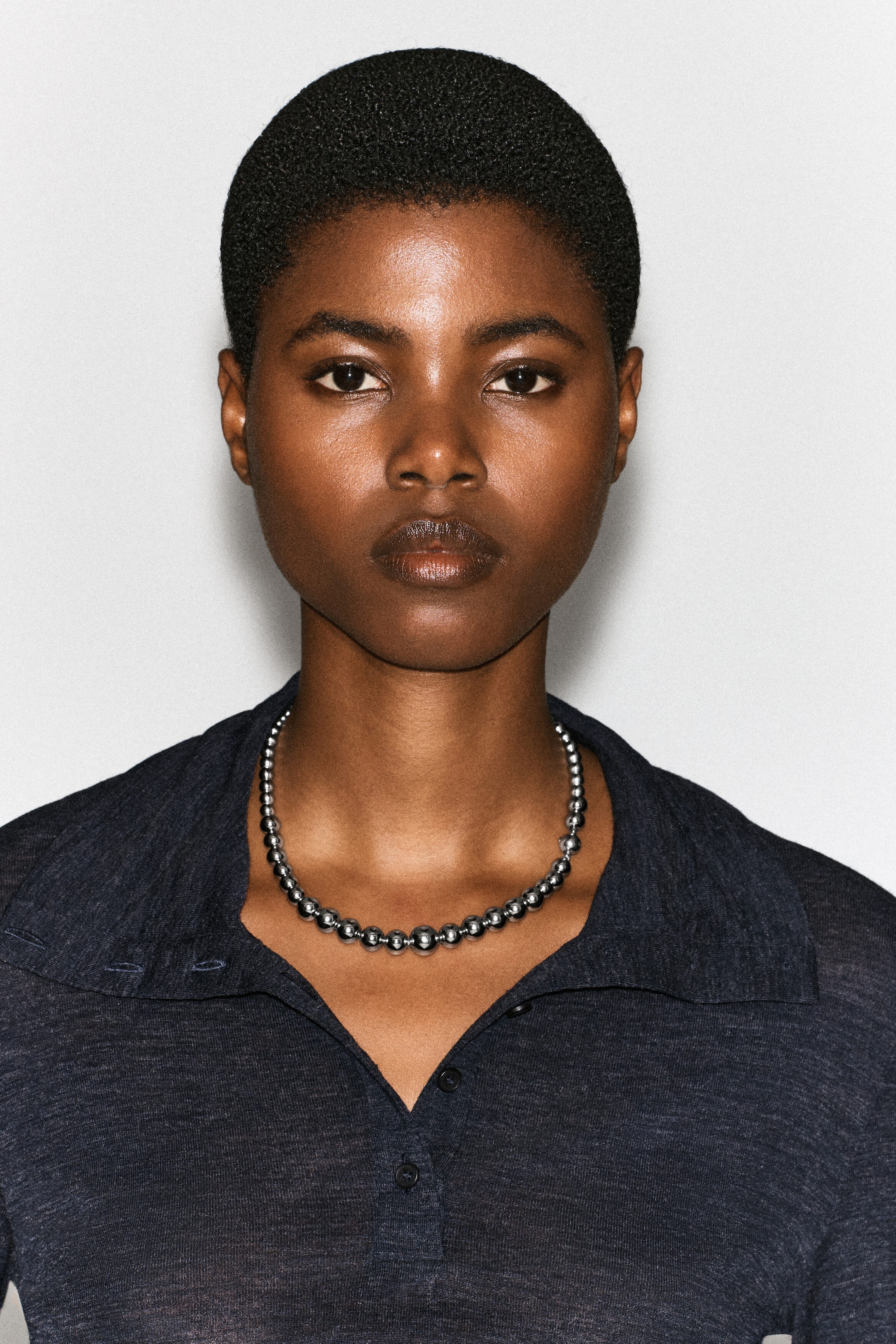 Un collier de perles composé de perles sphériques polies, couleur argentée, de tailles dégradées est porté par le mannequin. Le collier repose sur la clavicule, encadré par le col ouvert d’une chemise en maille gris foncé. Un collier de perles composé de perles sphériques polies, couleur argentée, de tailles dégradées est porté par le mannequin. Le collier repose sur la clavicule, encadré par le col ouvert d’une chemise en maille gris foncé.