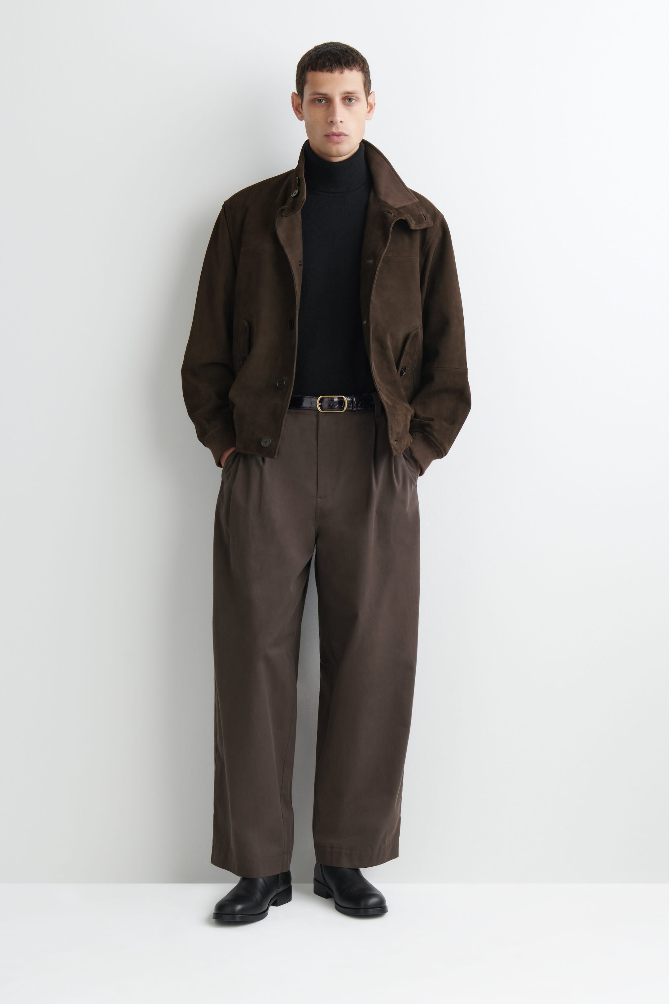 A dark brown suede jacket is displayed from the front, showcasing a relaxed fit, a wide collar, and ribbed cuffs. It is styled open over a black turtleneck sweater and paired with matching dark brown wide-leg pleated trousers. A dark brown suede jacket is displayed from the front, showcasing a relaxed fit, a wide collar, and ribbed cuffs. It is styled open over a black turtleneck sweater and paired with matching dark brown wide-leg pleated trousers.