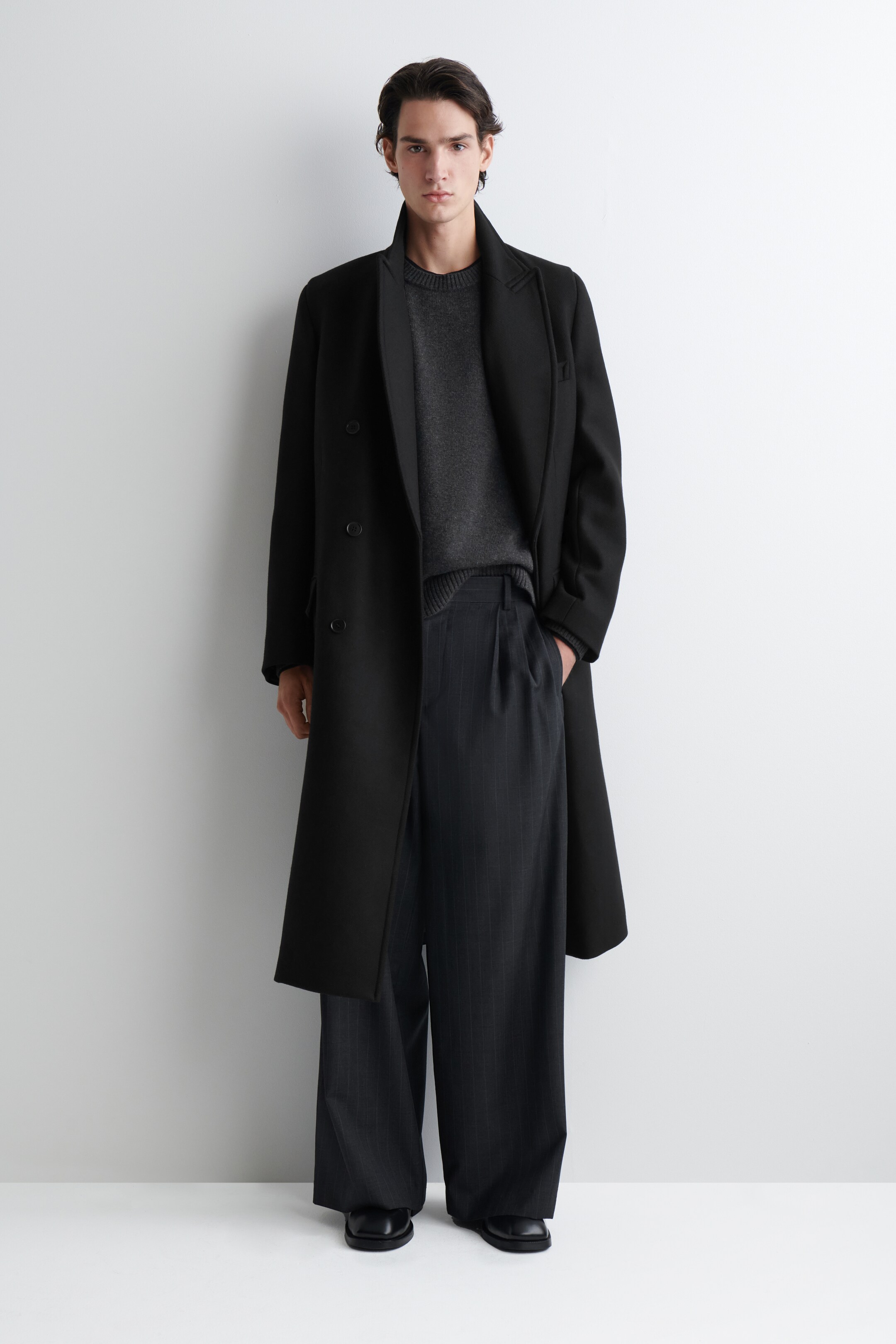 View larger image: A black double-breasted coat is being worn open, revealing its structured silhouette and notch lapels. Underneath, a dark grey crew-neck knit sweater is visible, along with wide-leg, dark grey pinstriped trousers featuring a single front pleat and black square-toed shoes.