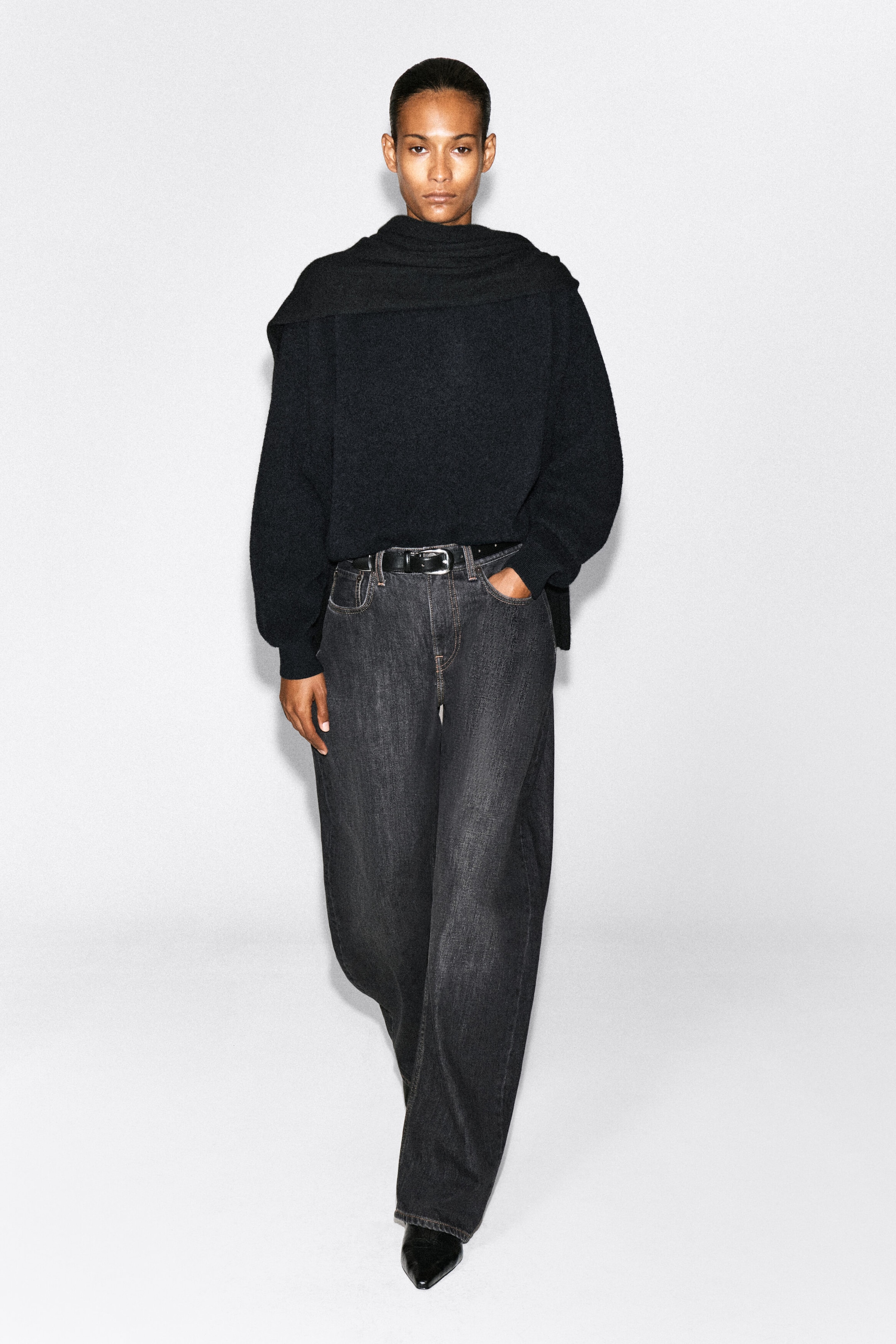 The model is wearing dark gray wide-leg jeans with a faded wash, standing with one hand resting in a front pocket. The jeans are styled with a black leather belt and paired with a black knit sweater featuring a draped scarf detail. The model is wearing dark gray wide-leg jeans with a faded wash, standing with one hand resting in a front pocket. The jeans are styled with a black leather belt and paired with a black knit sweater featuring a draped scarf detail.