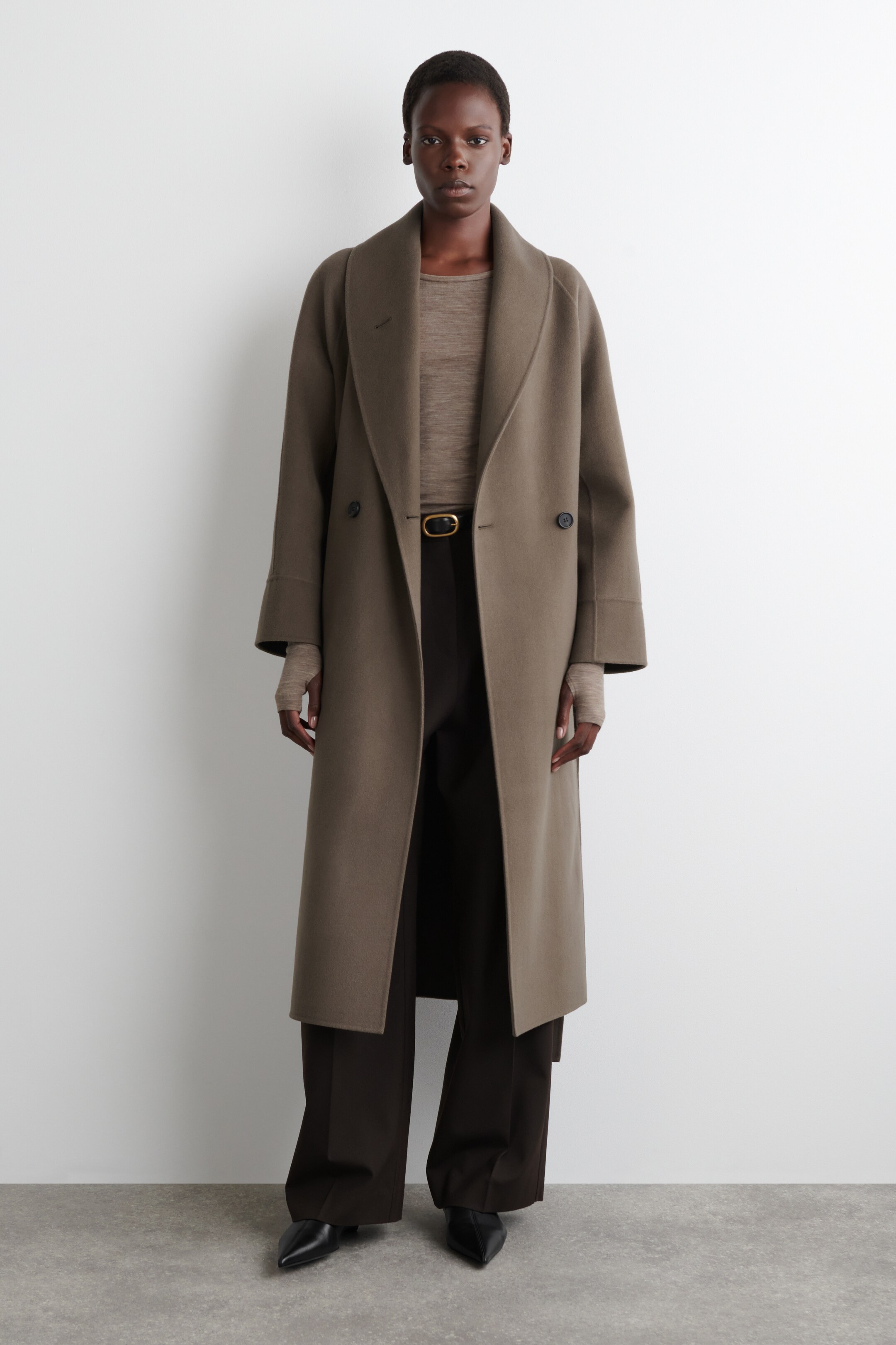 View larger image: A taupe long coat is being presented from the front, featuring a wide shawl collar and a double-breasted design with two visible buttons. The coat is styled open over a light brown top and dark trousers, showcasing its relaxed fit and long length.
