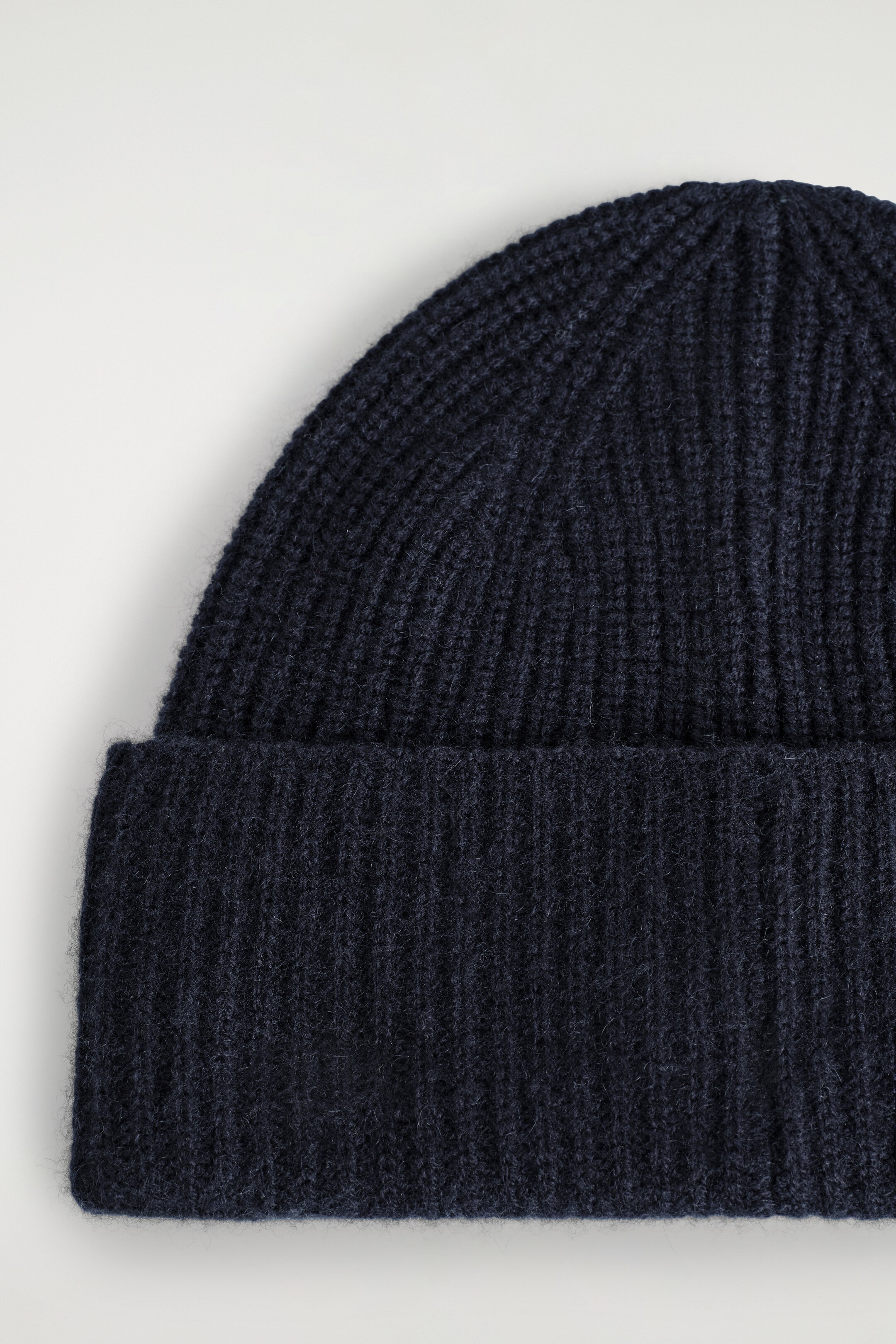View larger image: A dark blue beanie is being presented, showcasing its ribbed knit texture and a wide, folded cuff at the bottom.