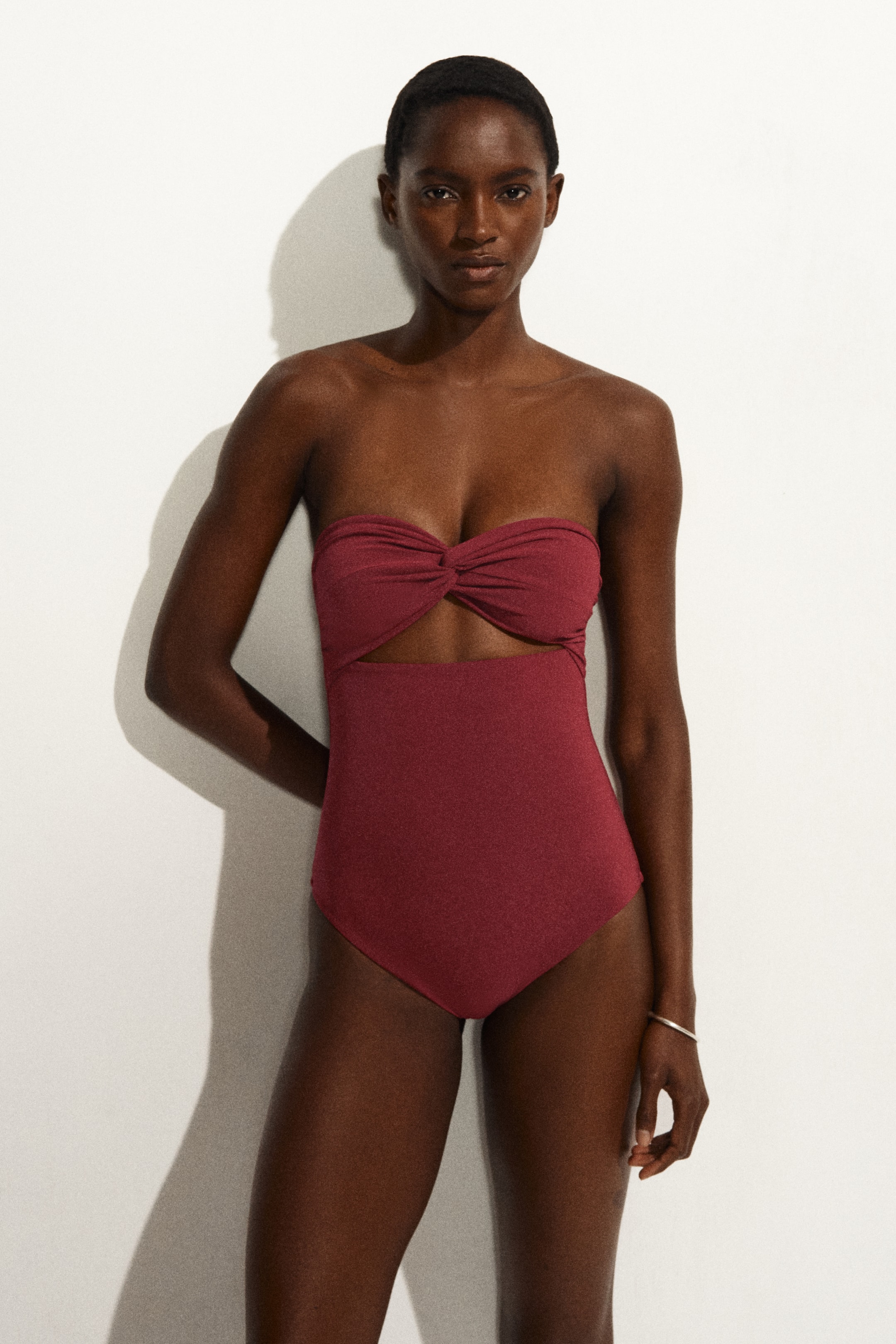 Ver imagen más grande: Un bañador rojo intenso de una sola pieza presenta un top bandeau sin tirantes con un detalle de nudo retorcido en el busto, creando una abertura en la parte inferior, y mostrando una línea de pierna de corte alto.