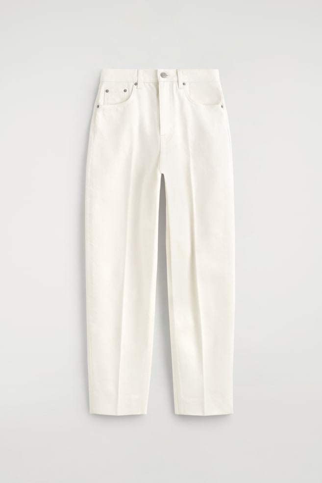 Un par de pantalones de color crema se presenta de frente, con una cintura que incluye trabillas para cinturón y un cierre de un solo botón. Se pueden ver dos bolsillos frontales, incluyendo un bolsillo para monedas, con remaches metálicos, y las perneras rectas terminan en dobladillos acabados. - 1