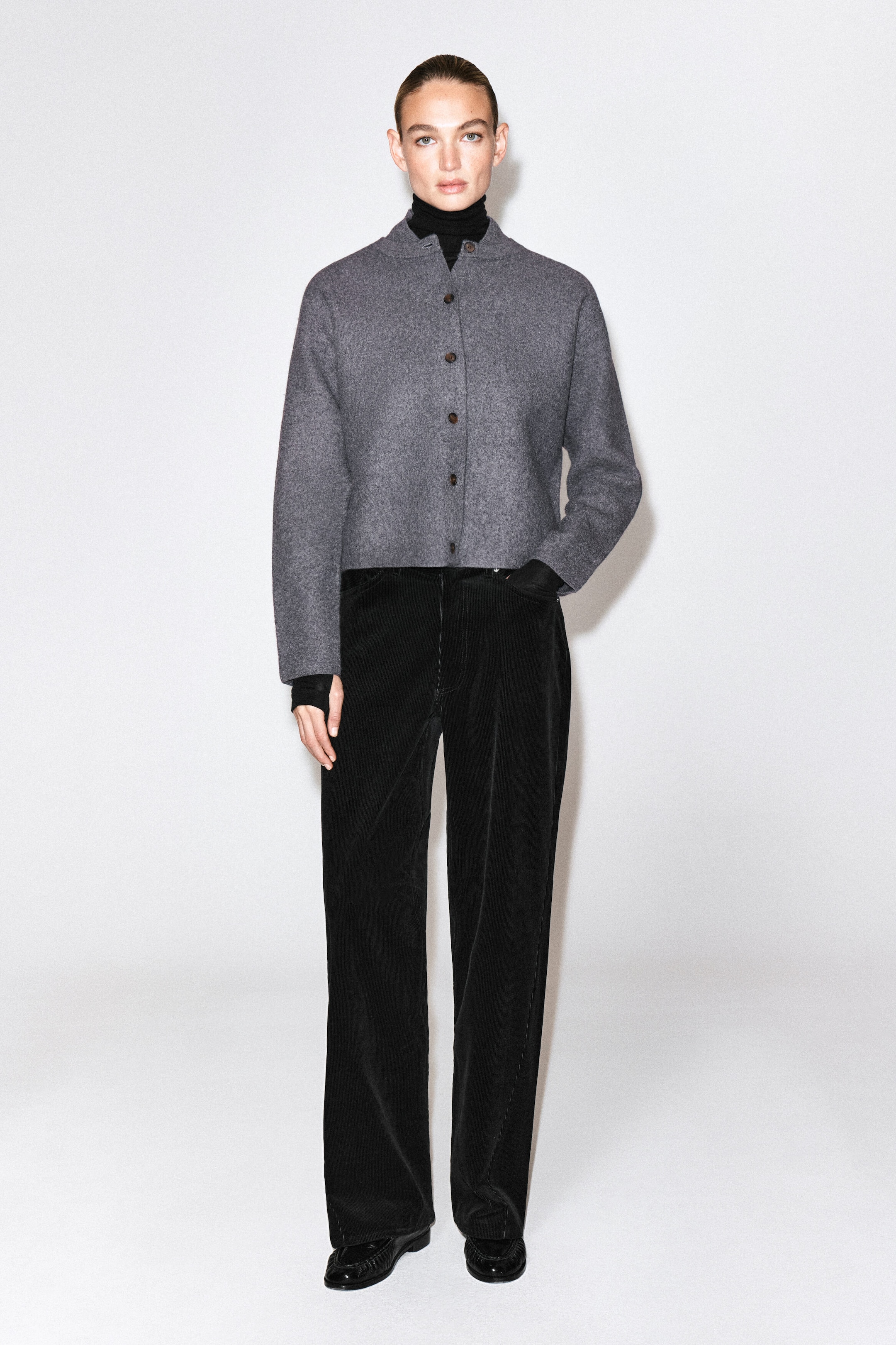 The model is wearing a gray boiled-wool cardigan featuring a button-front closure, a rounded silhouette, and a high collar layered over a black turtleneck. The cardigan is styled with black wide-leg trousers. The model is wearing a gray boiled-wool cardigan featuring a button-front closure, a rounded silhouette, and a high collar layered over a black turtleneck. The cardigan is styled with black wide-leg trousers.