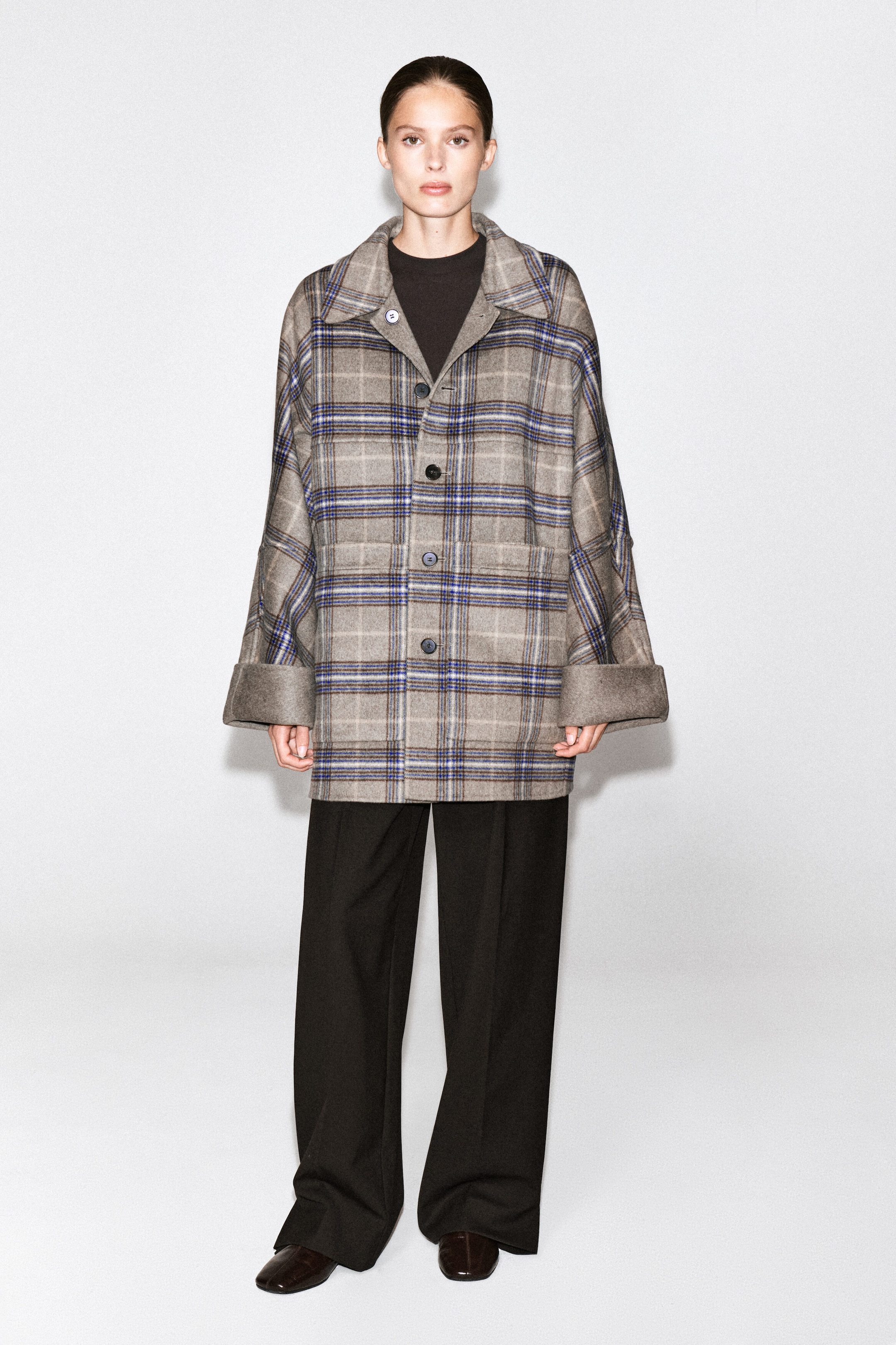 The model is wearing a reversible check coat featuring a button-front closure, a classic plaid pattern in gray, brown, and blue, and wide sleeves with folded cuffs that reveal a solid beige lining. The coat is styled as a layer over a black high-neck top and relaxed, wide-leg trousers.