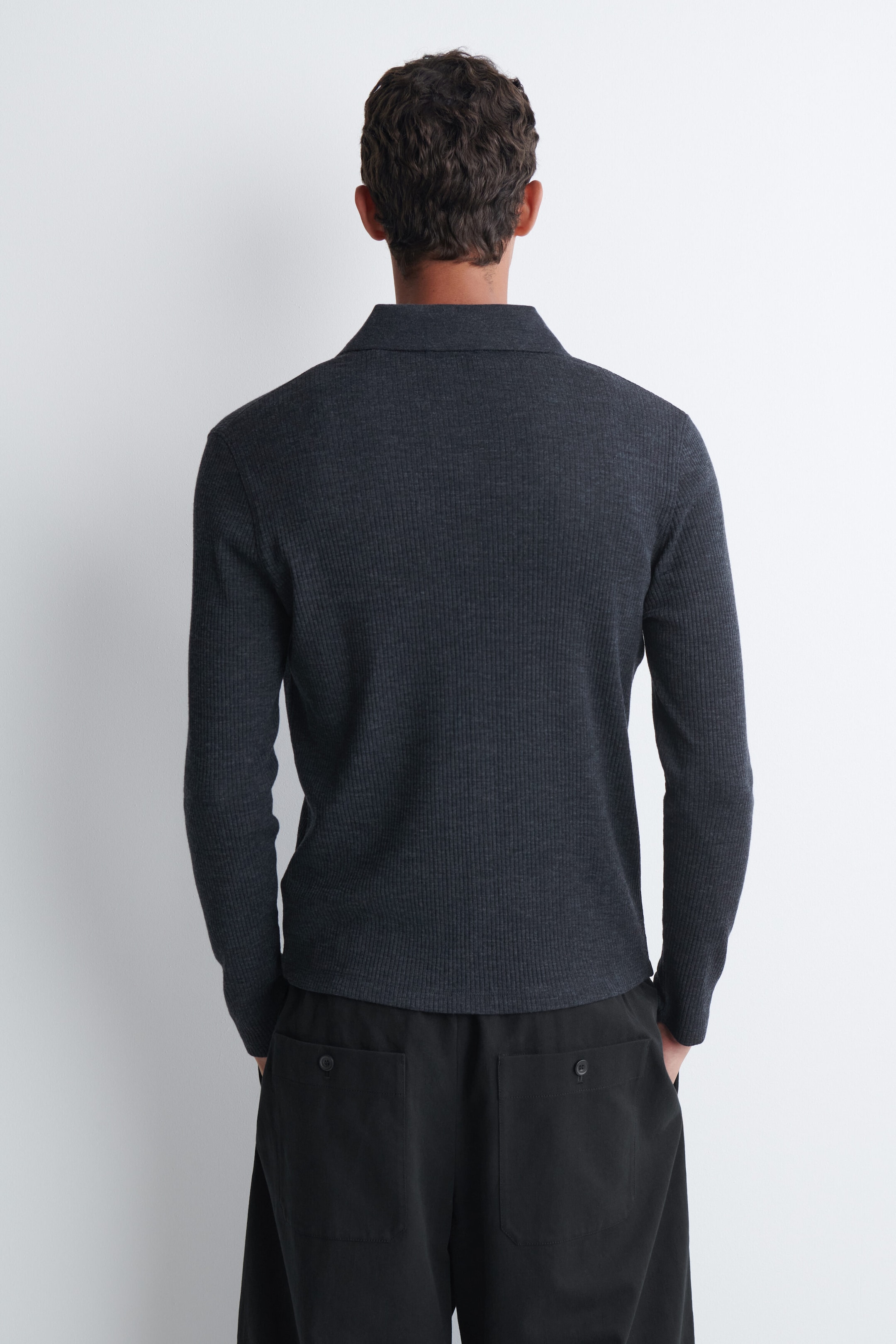 A dark grey ribbed-knit long-sleeve polo shirt is being worn by the model, shown from the back. The shirt features a classic collar and is paired with black trousers.