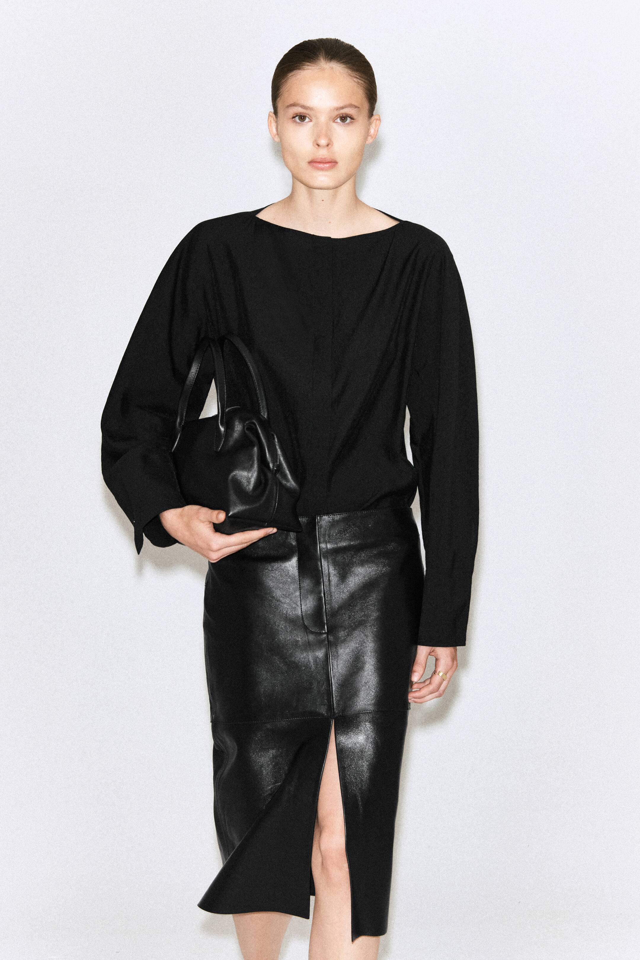 Vezi imaginea mai mare: Modelul poartă o bluză neagră cu un decolteu bărcuță curat și mâneci lungi. Bluza are un drapaj fluid și o siluetă sculpturală, fiind asortată cu o fustă din piele neagră.