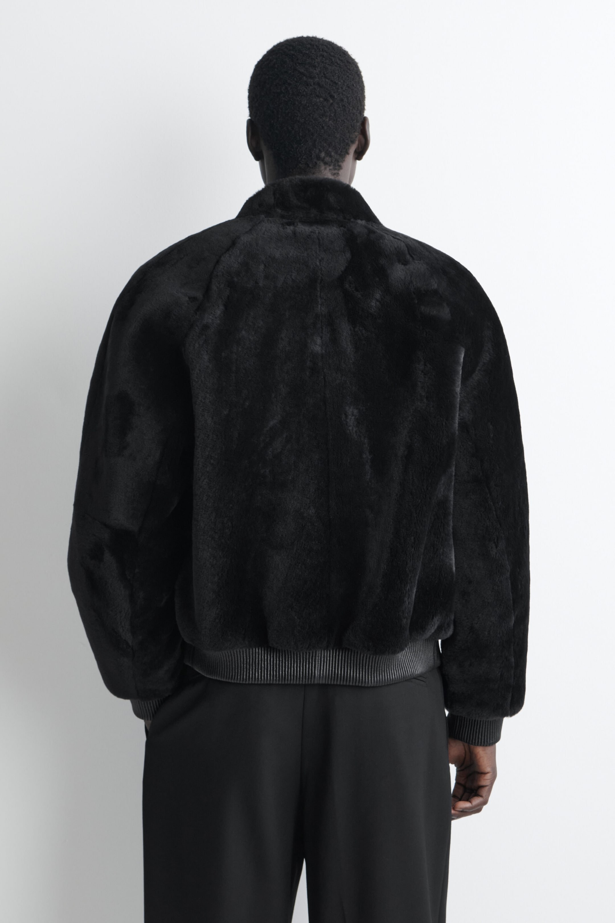 A black padded shearling bomber jacket is shown from the back, showcasing its plush, textured finish and structured, voluminous silhouette. The jacket features a high collar, raglan-style shoulder seams, and a ribbed hem and cuffs.