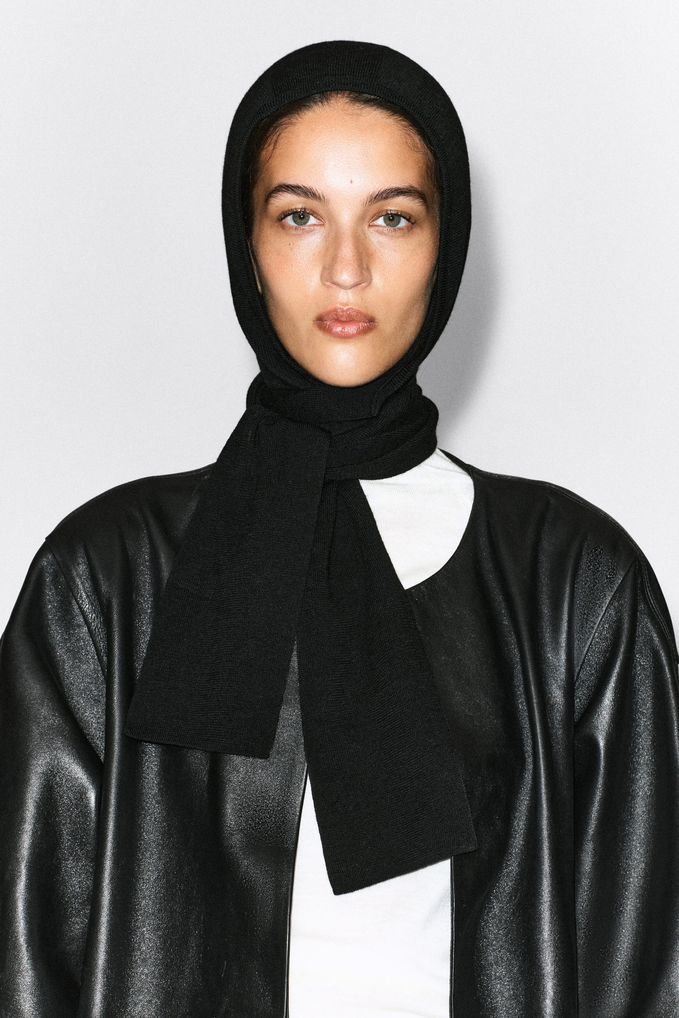 A black knit hooded scarf is being worn by the model, covering the head and wrapping around the neck with the ends hanging down the front. The scarf is styled over a white top and a black leather jacket. A black knit hooded scarf is being worn by the model, covering the head and wrapping around the neck with the ends hanging down the front. The scarf is styled over a white top and a black leather jacket.