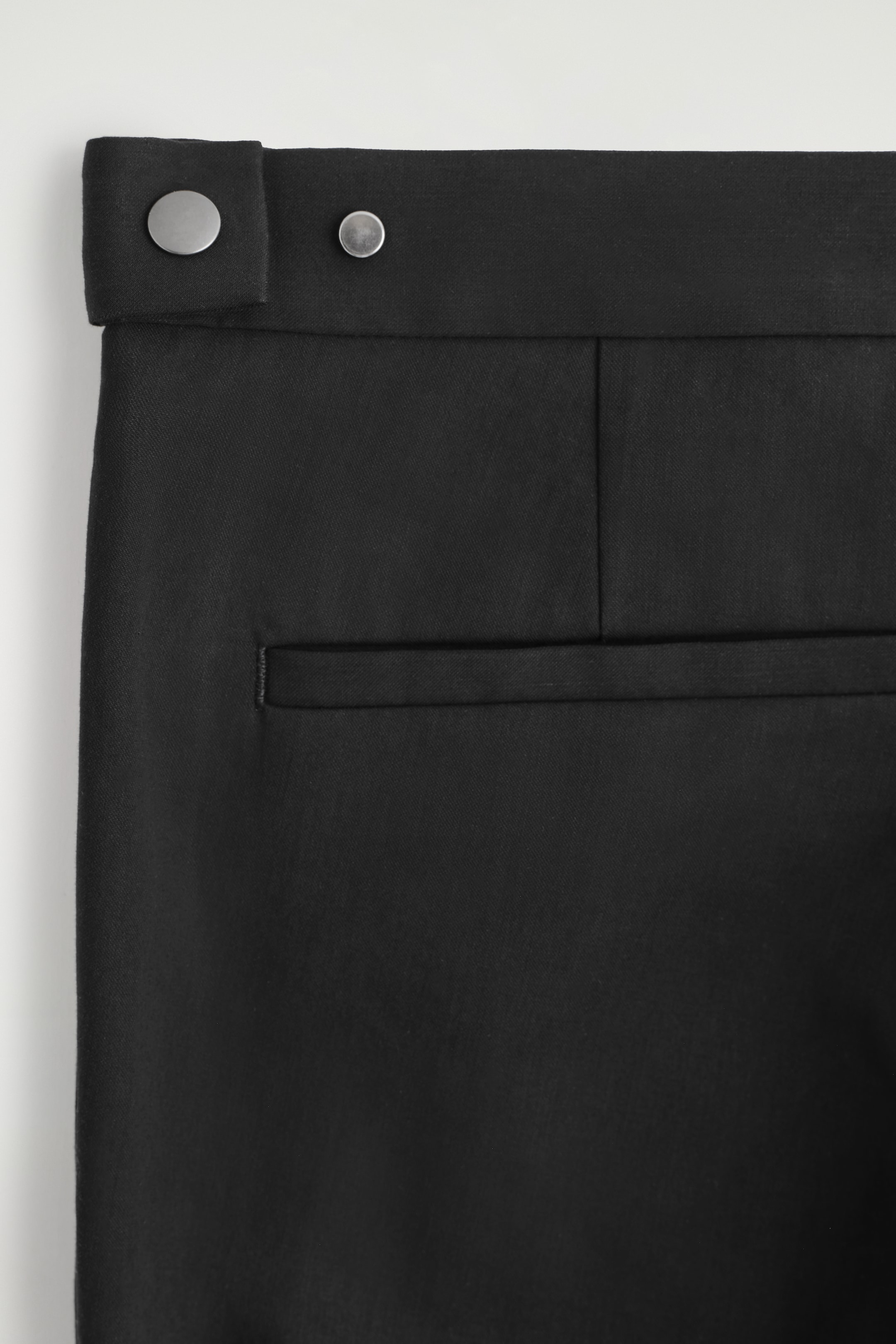 View larger image: The back of black trousers is shown, featuring a wide waistband with two silver-toned press studs on the left side. A horizontal welt pocket is positioned on the right side below the waistband.