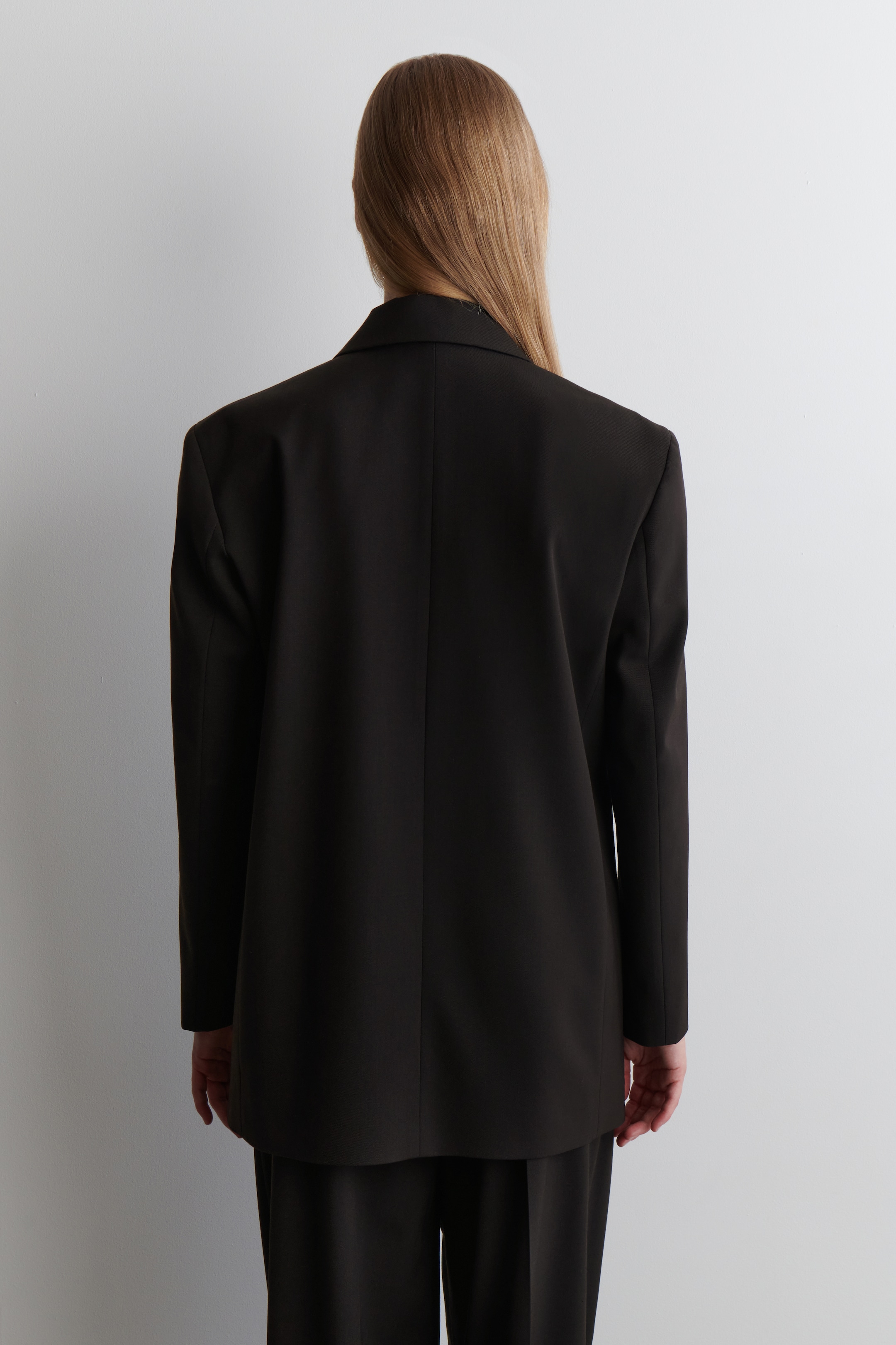 Visualizza immagine più grande: Un blazer nero viene mostrato da dietro, caratterizzato da una vestibilità rilassata con spalle strutturate e una cucitura centrale posteriore, indossato sopra pantaloni neri coordinati.