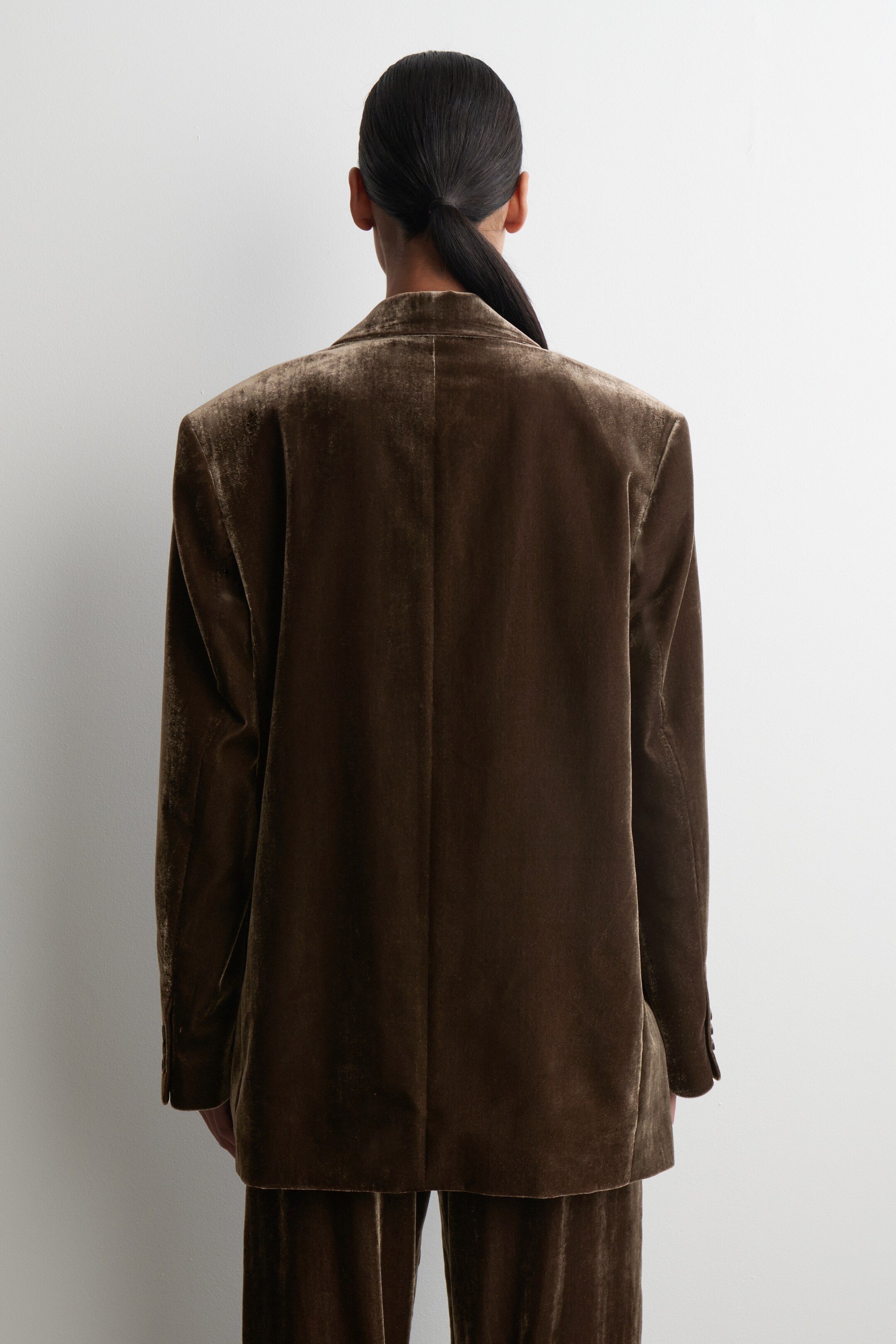 Agrandir l'image: Un blazer en velours marron est présenté de dos, mettant en valeur sa coupe décontractée et sa texture douce. Il présente une couture centrale visible au dos. Un pantalon en velours assorti est également porté, apparaissant sous le blazer.