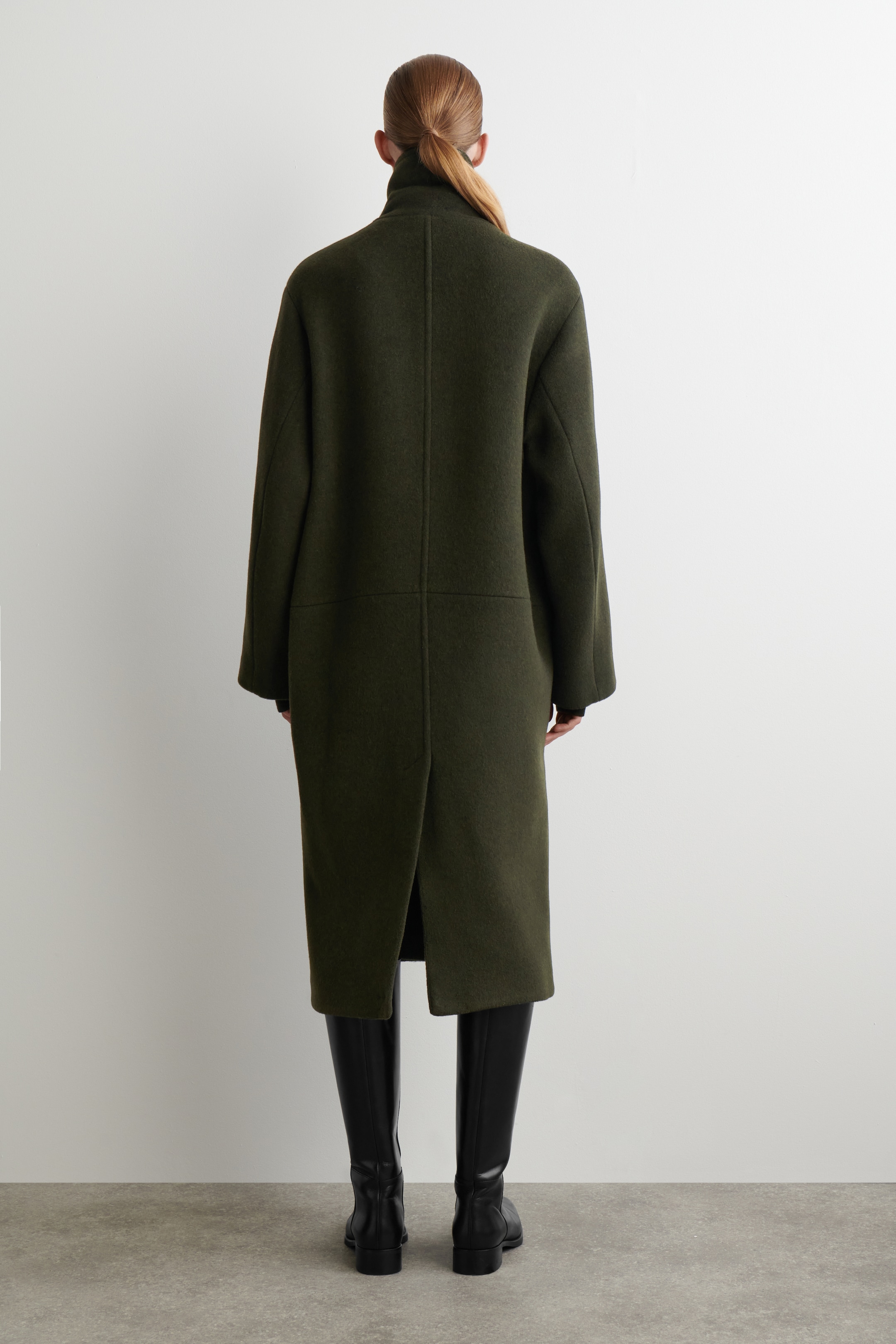 Visualizza immagine più grande: Un cappotto verde oliva viene presentato dalla vista posteriore, caratterizzato da un colletto alto e una cucitura centrale posteriore prominente. Il cappotto è progettato con una vestibilità rilassata e dritta, maniche lunghe e uno spacco centrale posteriore profondo, che si estende fino a metà polpaccio sopra stivali neri alti fino al ginocchio.