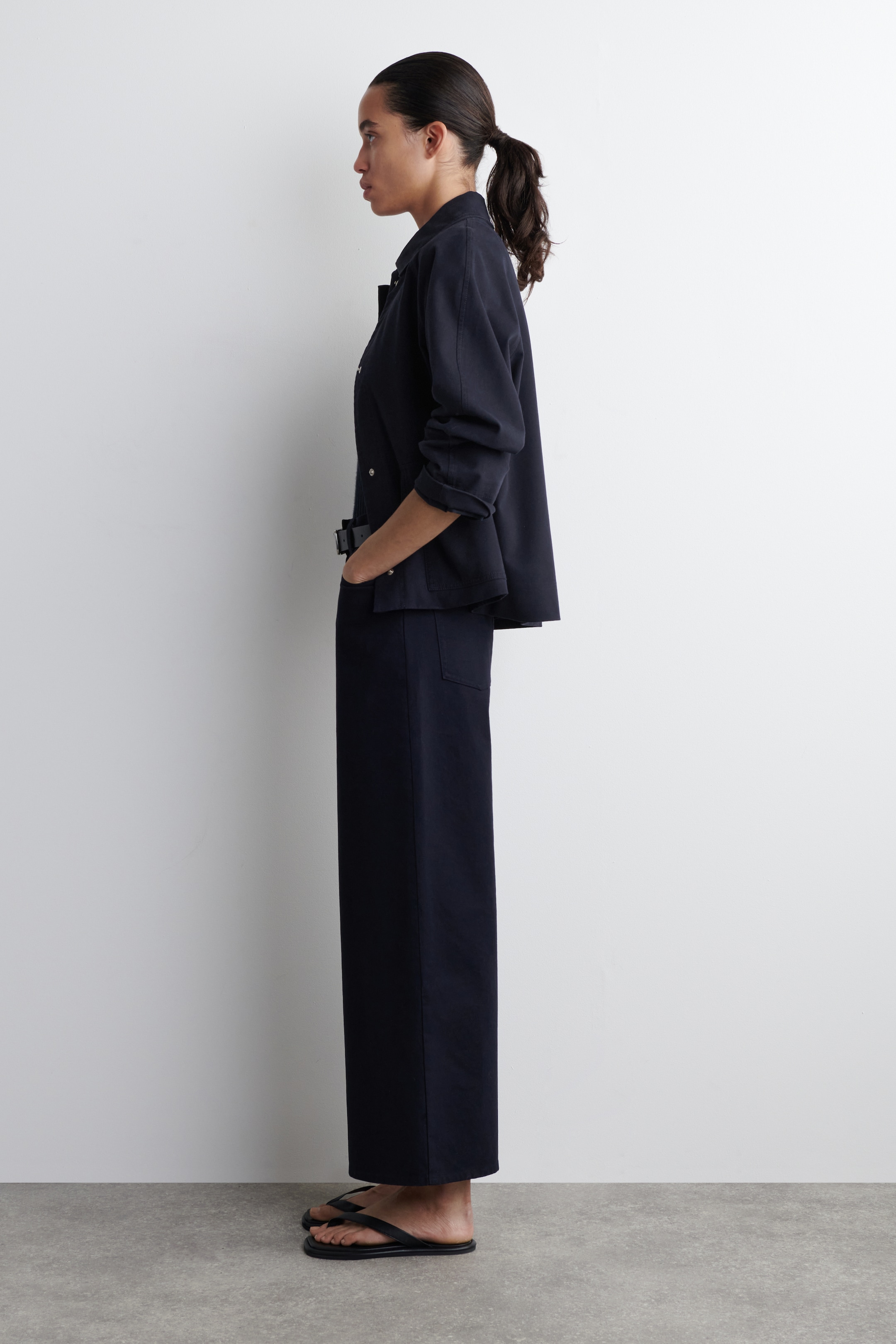 Größeres Bild anzeigen: Ein marineblaues Outfit wird aus einer Seitenansicht präsentiert, das eine locker sitzende Langarm-Hemdjacke mit einem Standardkragen, sichtbaren Vorderknöpfen und bis zu den Unterarmen hochgekrempelten Ärmeln zeigt. Ein schwarzer Schnallengurt zieht die Taille der Jacke zusammen und vervollständigt den Look mit passenden, weit geschnittenen Hosen, die bis zu den Knöcheln reichen, und schwarzen Flip-Flops, die getragen werden.