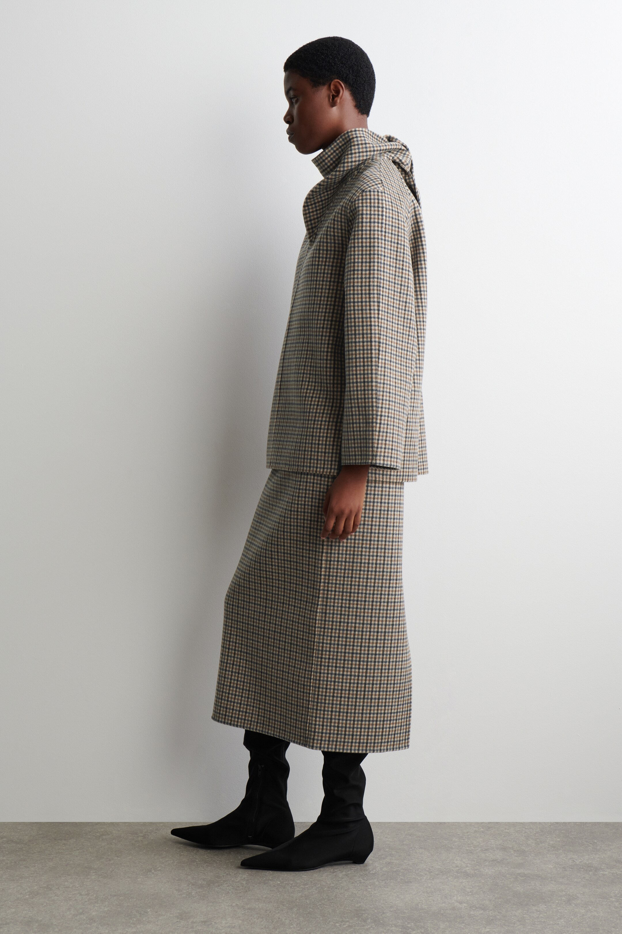 Ver imagen más grande: Un conjunto de dos piezas, que presenta una chaqueta con un cuello alto y una falda midi a juego, se presenta en un patrón de cuadros pequeños en marrón, negro y blanco, visto desde el perfil izquierdo. Se llevan botas negras de punta afilada con el conjunto.