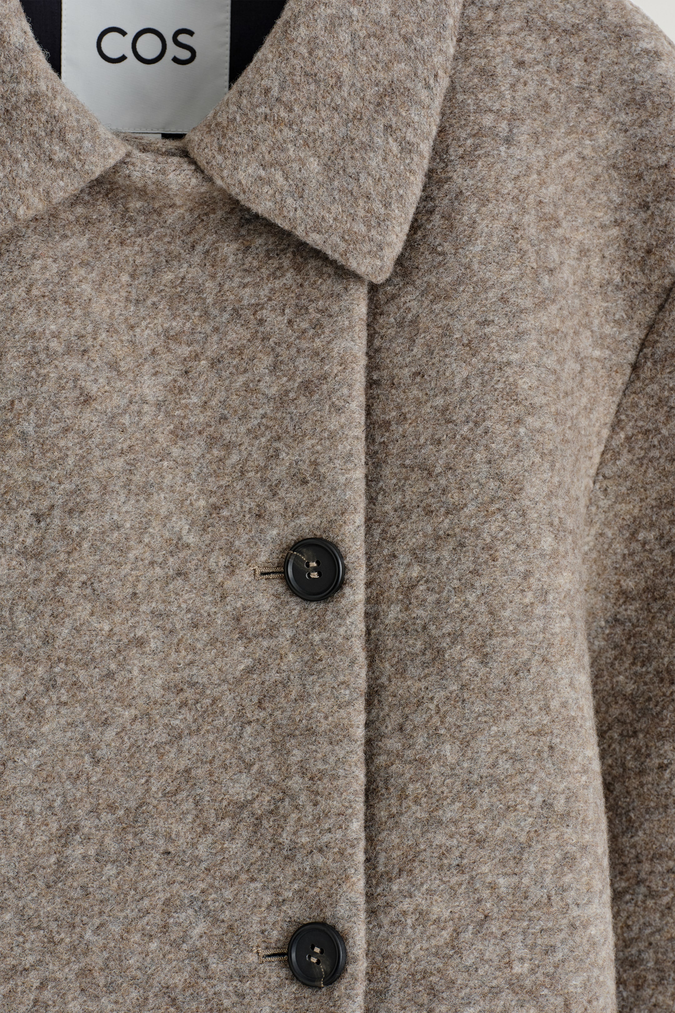 Ver imagen más grande: Una vista de cerca muestra la parte delantera de una chaqueta de melange marrón claro, hecha de un tejido texturizado y ligeramente peludo, y con un cuello ancho y plano. Dos botones negros están alineados verticalmente y posicionados descentrados hacia la izquierda de una costura central, formando el cierre frontal, con una etiqueta blanca de la marca COS visible detrás del cuello.