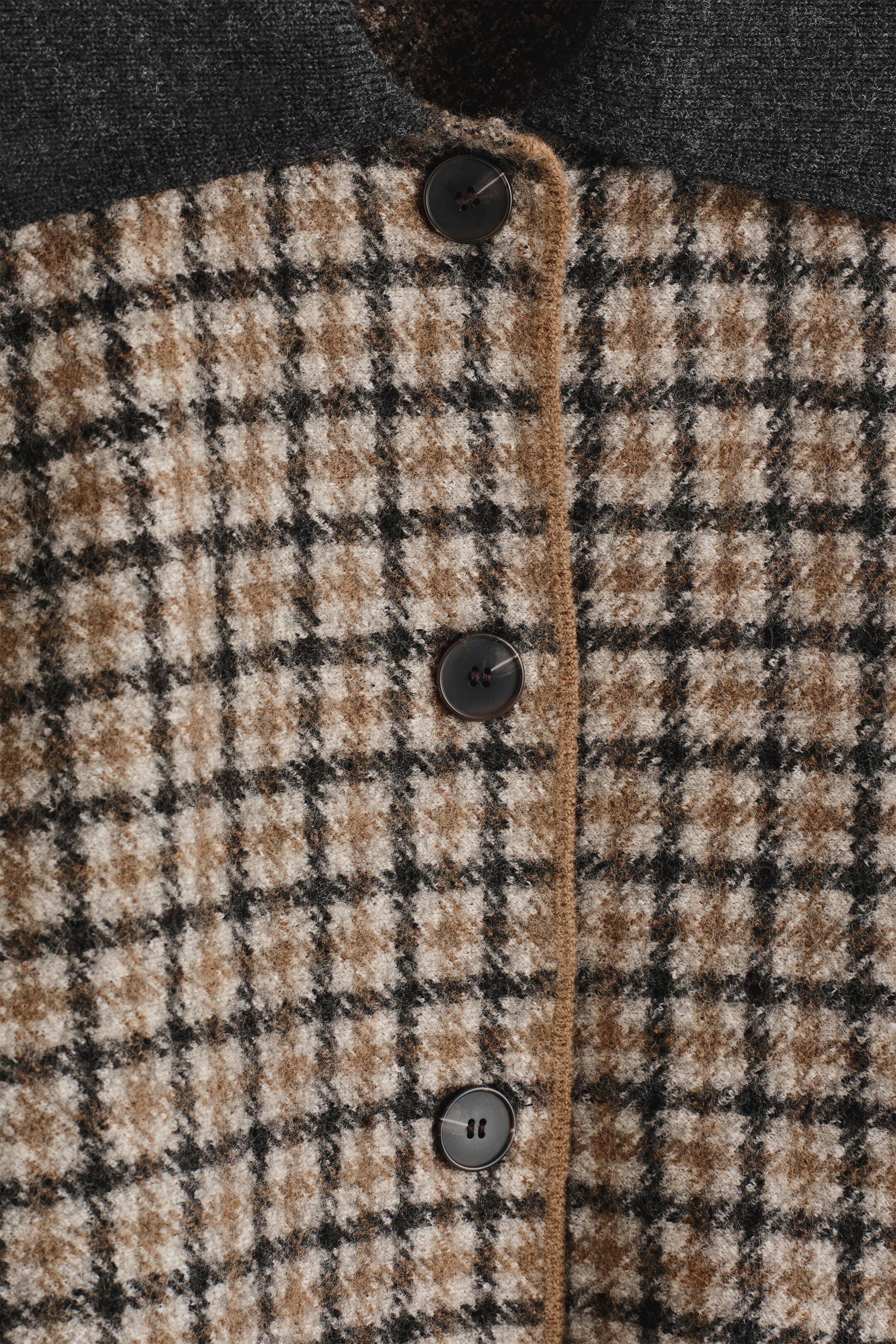 View larger image: A jacket as FRONT is displaying a brown, black, and cream plaid pattern, with a dark grey ribbed collar visible at the top. Three dark, round buttons are fastening the jacket down the center, alongside a narrow brown trim on the placket.