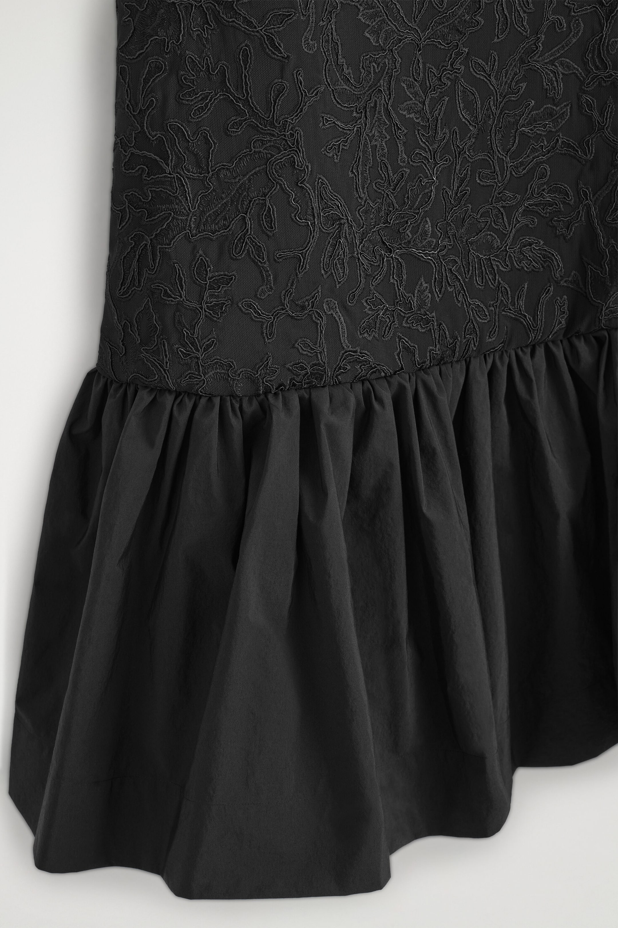 A close-up of a black dress showing the transition from a bodice with intricate tonal floral embroidery on a mesh background to a gathered, voluminous poplin skirt. A close-up of a black dress showing the transition from a bodice with intricate tonal floral embroidery on a mesh background to a gathered, voluminous poplin skirt.