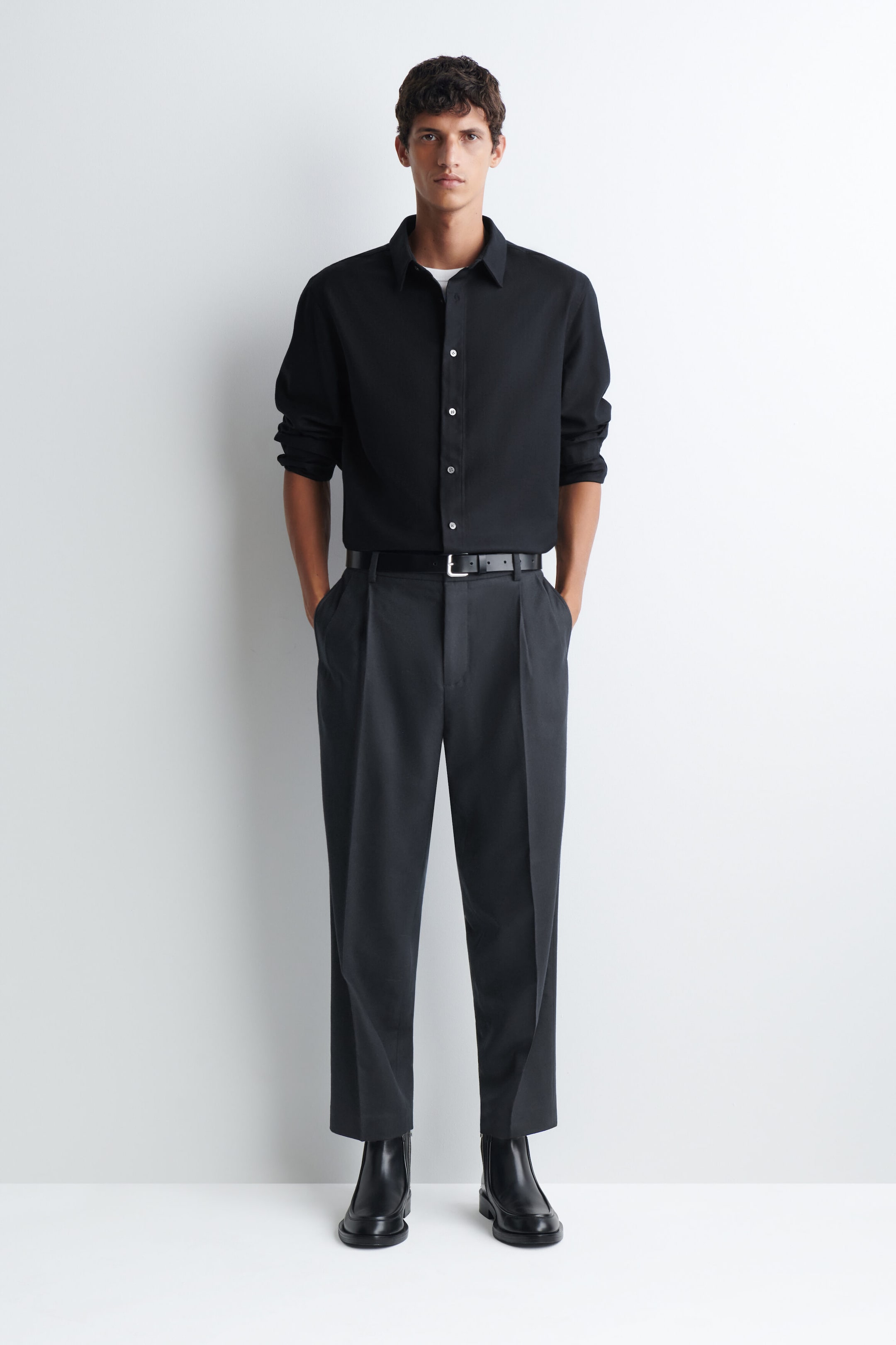 The model is wearing dark gray pleated trousers, paired with a tucked-in black long-sleeve shirt and a black leather belt. The trousers feature a relaxed fit with a sharp front crease and are styled with black leather boots. The model is wearing dark gray pleated trousers, paired with a tucked-in black long-sleeve shirt and a black leather belt. The trousers feature a relaxed fit with a sharp front crease and are styled with black leather boots.