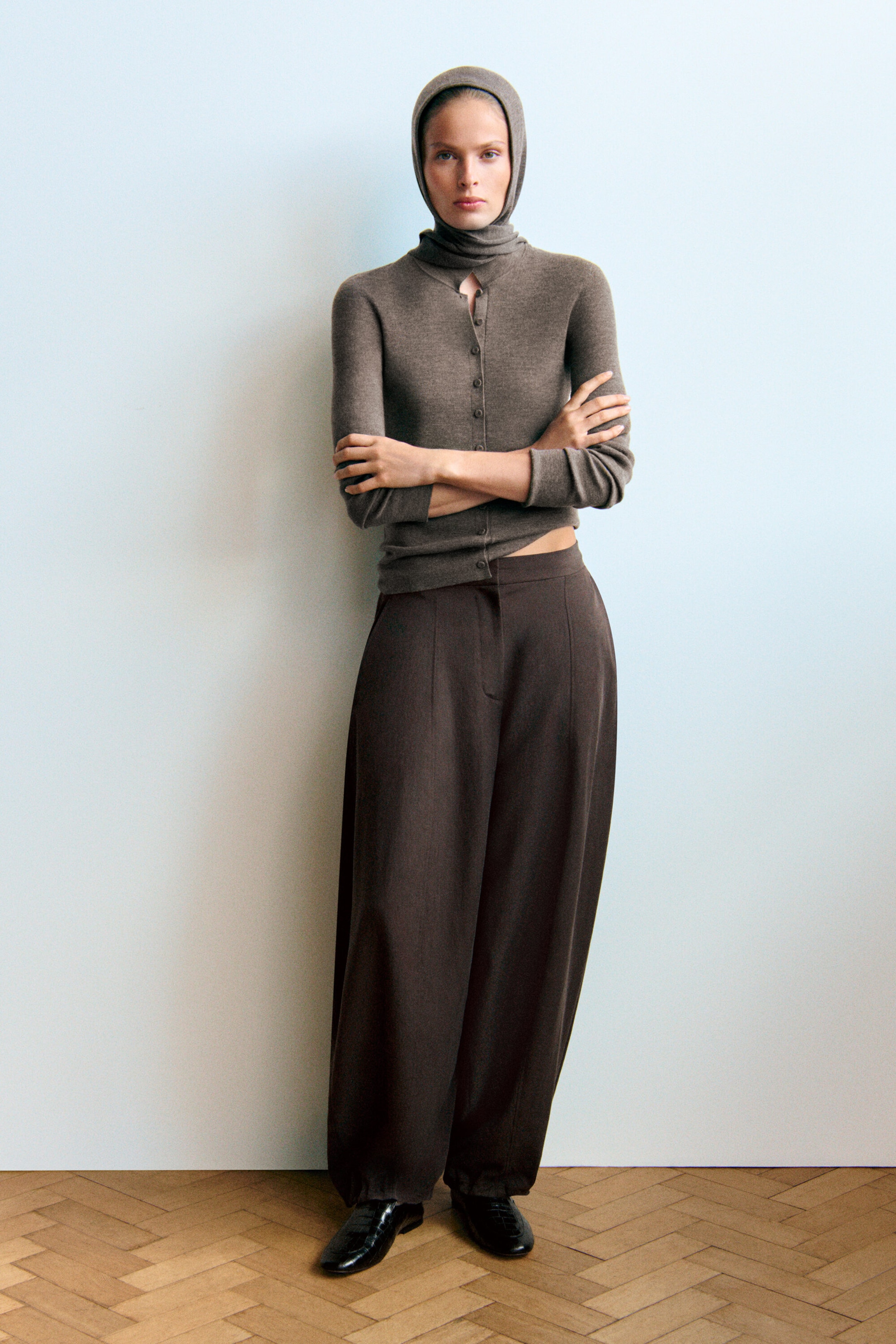 A model is standing front-facing, wearing a slim-fit taupe-brown merino wool knitted shirt with fabric-covered buttons and a matching merino hooded scarf worn over the head. The outfit is completed with high-waisted dark brown balloon trousers featuring a wide-leg silhouette. A model is standing front-facing, wearing a slim-fit taupe-brown merino wool knitted shirt with fabric-covered buttons and a matching merino hooded scarf worn over the head. The outfit is completed with high-waisted dark brown balloon trousers featuring a wide-leg silhouette.