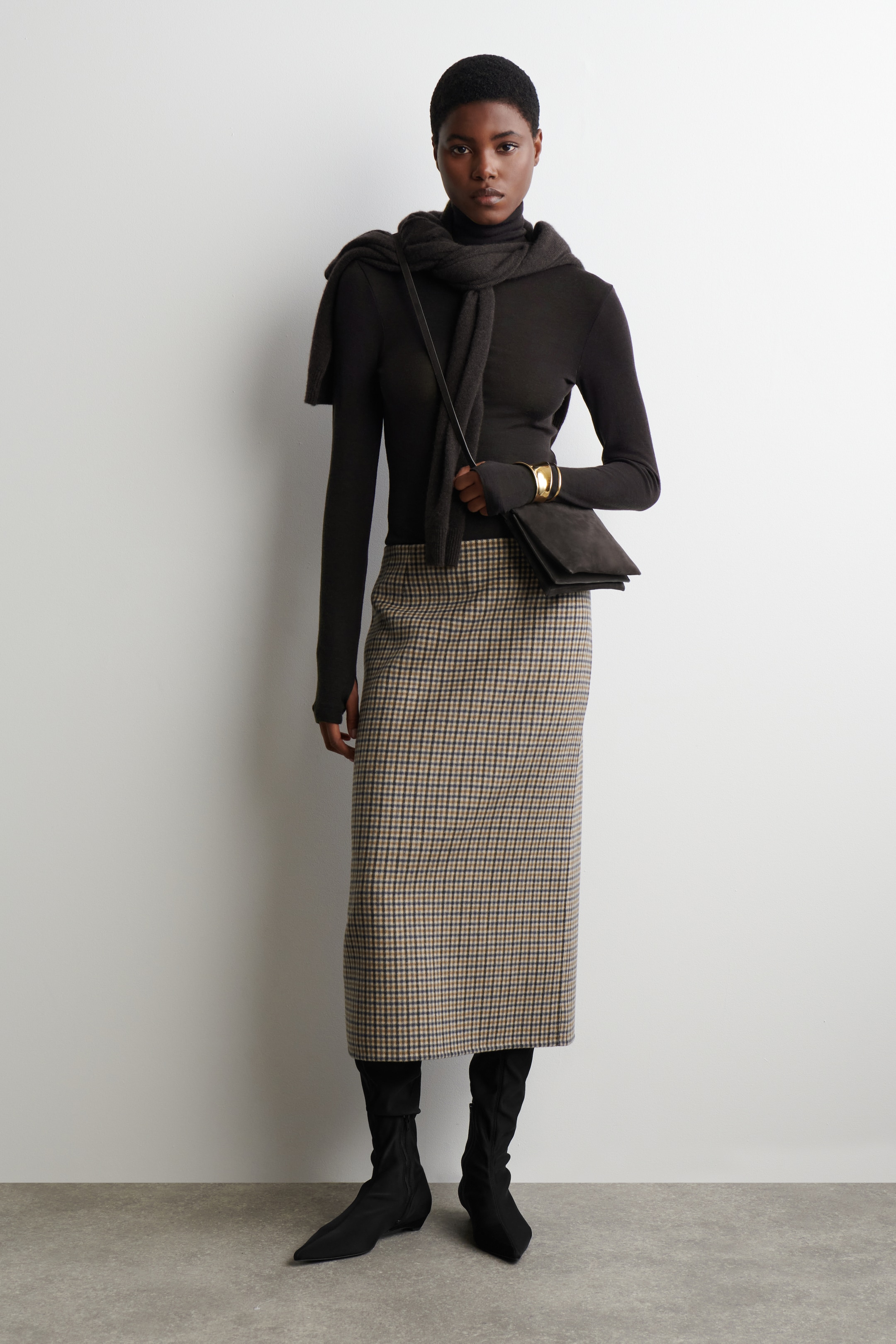 Agrandir l'image: Une tenue est présentée sur un mannequin, mettant en avant un haut à col roulé à manches longues et ajusté de couleur sombre avec des trous pour les pouces, stylisé avec une écharpe sombre assortie drapée autour de son cou et de ses épaules. Elle porte une jupe droite de longueur midi avec un petit motif à carreaux en beige et tons sombres. Le look est complété par des bottines noires à bout pointu, un petit sac bandoulière sombre et un large bracelet manchette en or.