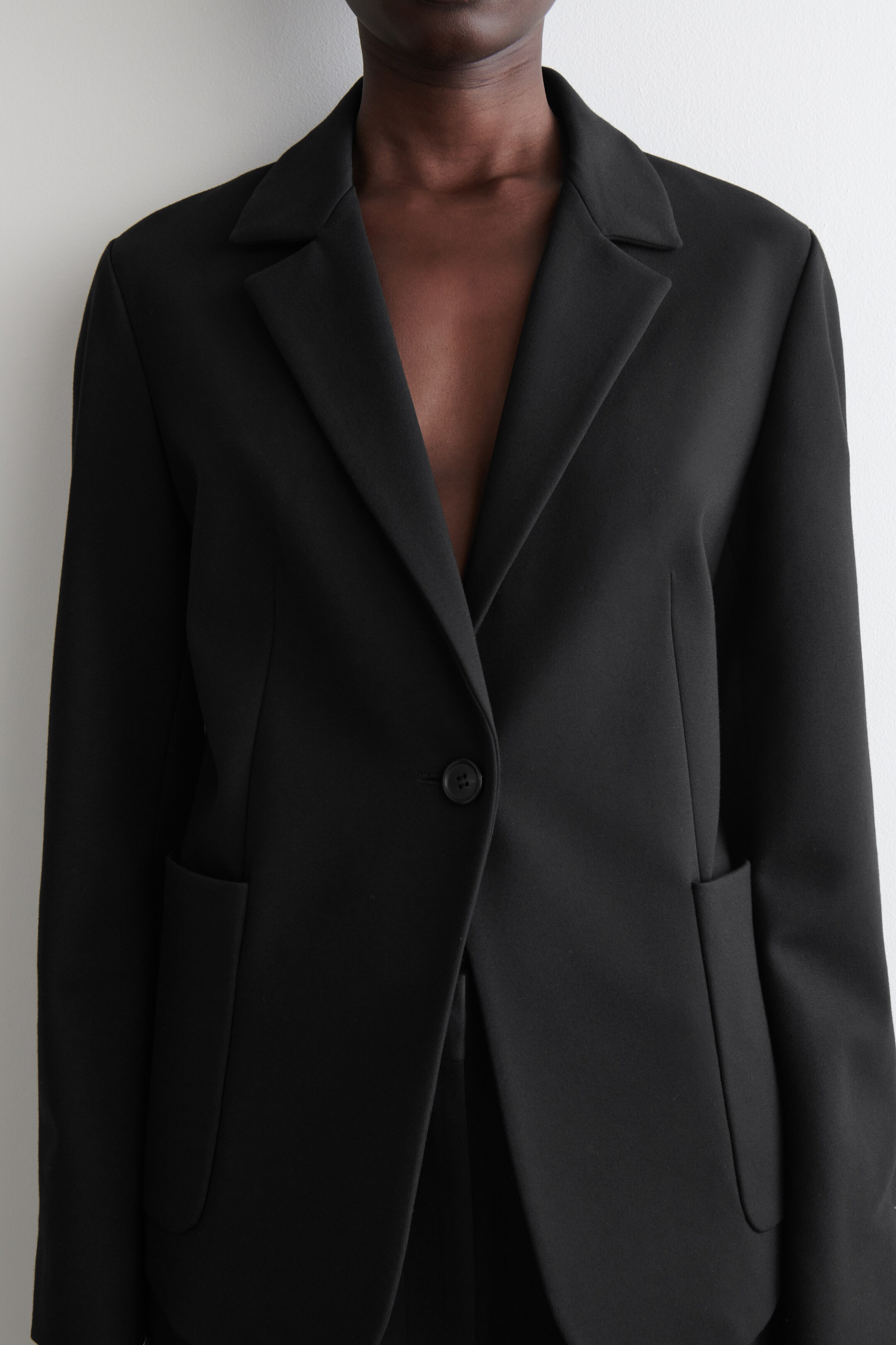 Zobrazit větší obrázek: Černé sako s jedním řadovým zapínáním je nošeno, mírně nakloněné doprava. Má široké klopy s výřezem, jednořadové zapínání na knoflík a dvě velké našité kapsy na dolní přední části.