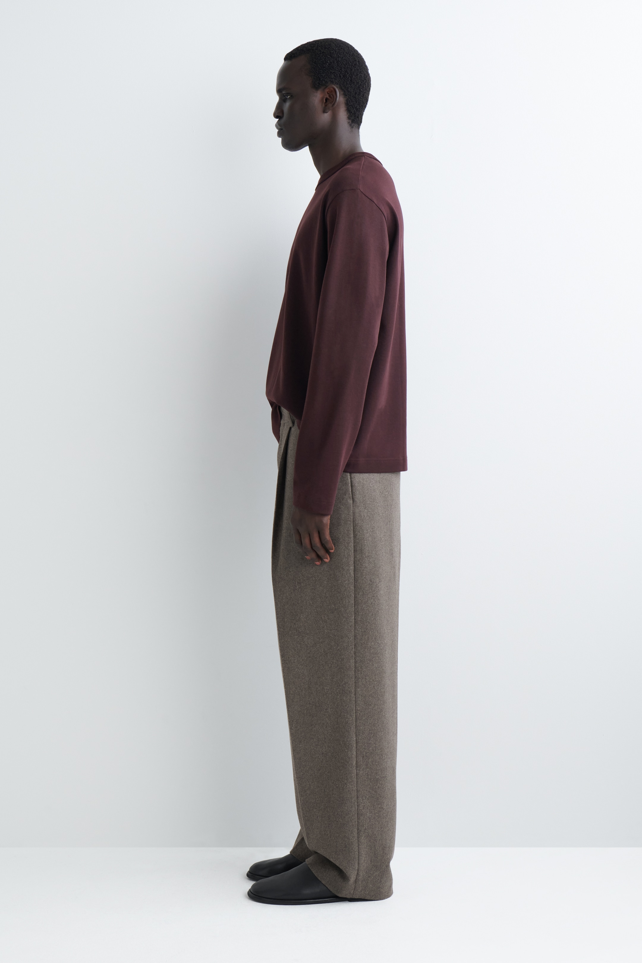 Visualizza immagine più grande: Una maglia a maniche lunghe con scollo rotondo di colore rosso scuro e vestibilità rilassata è abbinata a pantaloni a gamba larga e pieghe di colore marrone scuro, mostrati da un'angolazione laterale.