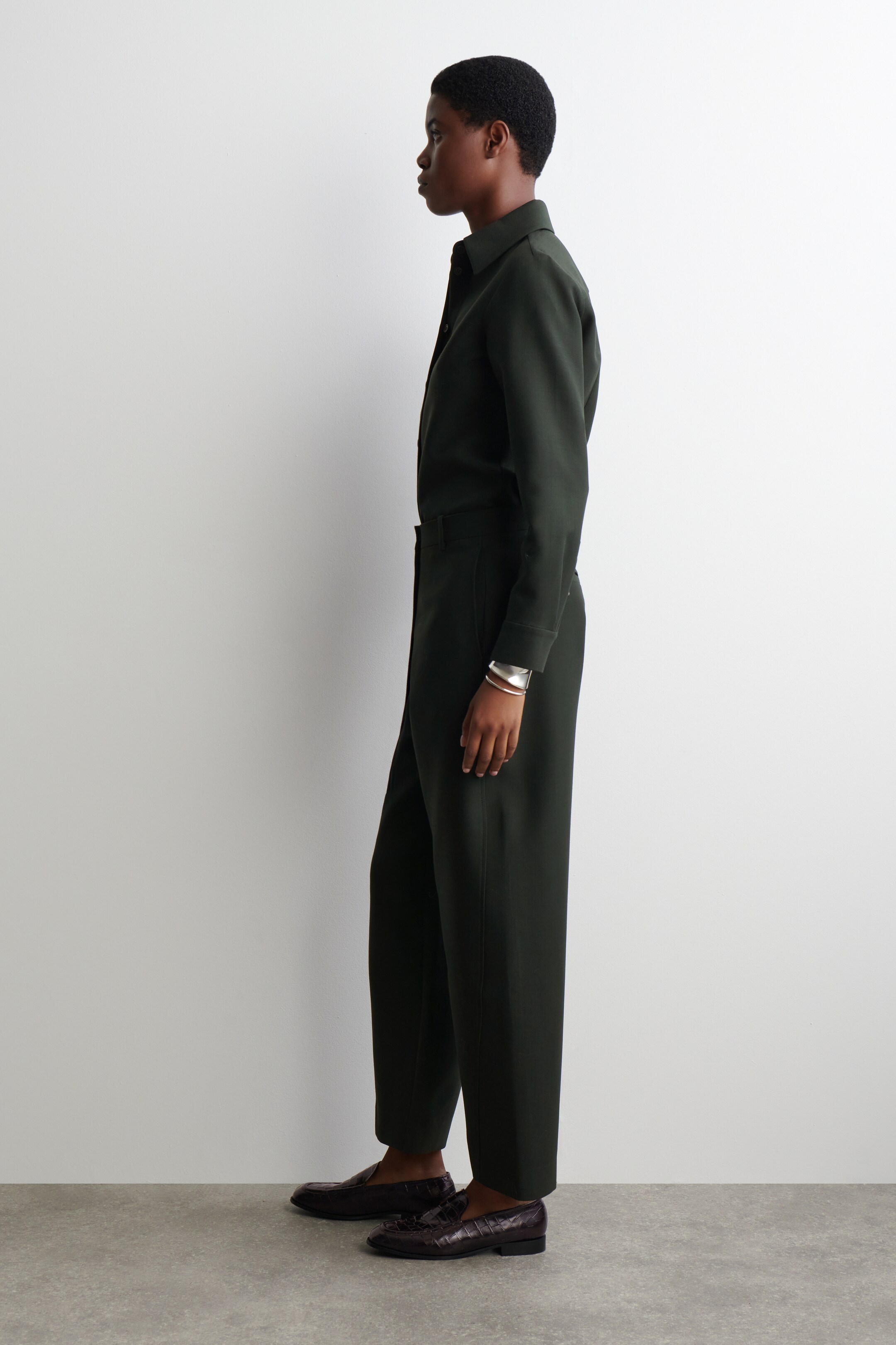 Agrandir l'image: Une chemise à col vert foncé avec des manches longues est assortie à un pantalon droit à coupe décontractée de la même couleur. L'ensemble est vu de côté gauche, complété par des mocassins embossés en crocodile marron foncé et un bracelet manchette en argent.