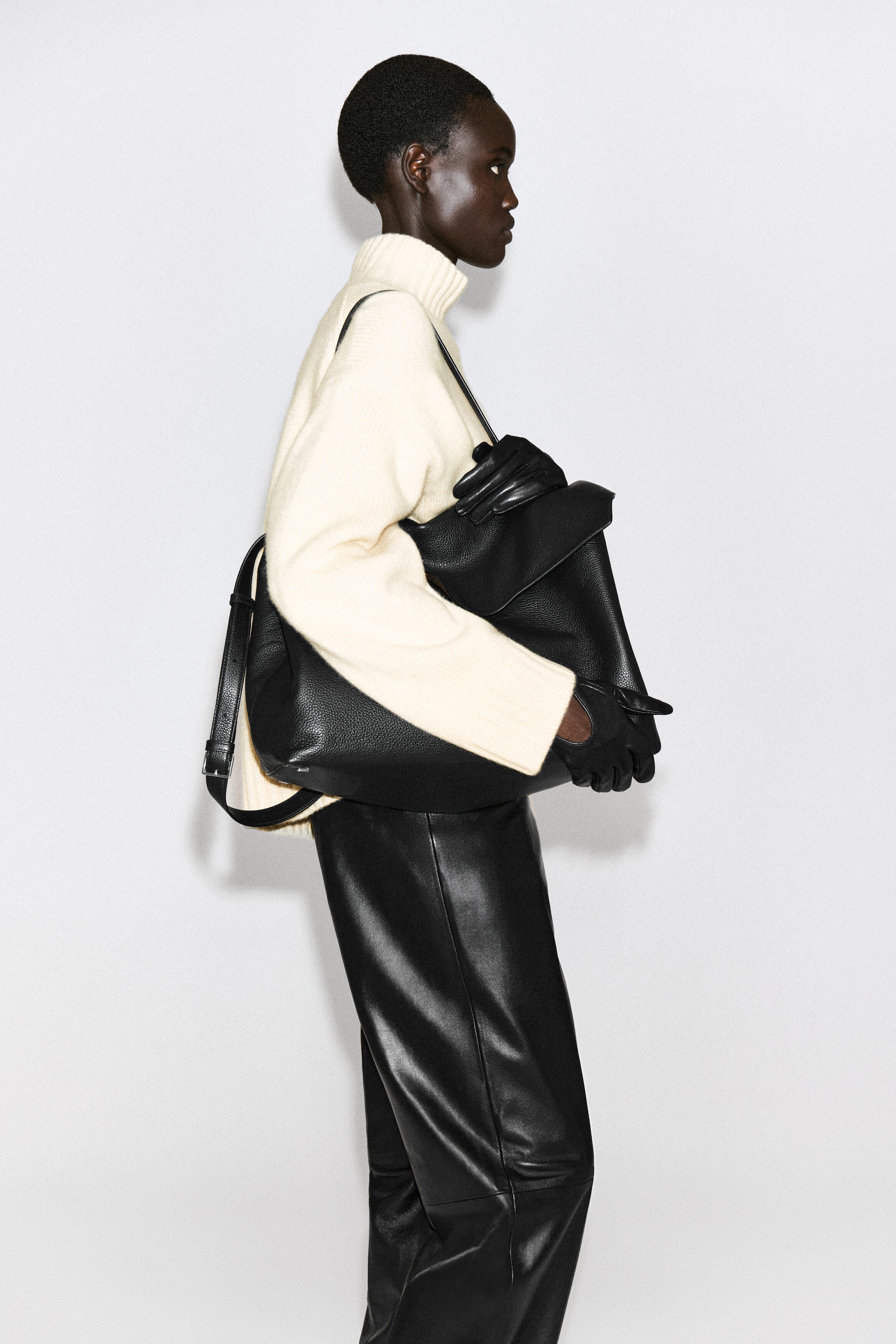 Black leather driving gloves are being worn by a model facing right, with the snug-fitting gloves extending from the wide cuffs of a cream-coloured knit sweater. The gloved hands are holding a large black pebbled-leather bag, showcasing the gloves' smooth finish and flexible design. Black leather driving gloves are being worn by a model facing right, with the snug-fitting gloves extending from the wide cuffs of a cream-coloured knit sweater. The gloved hands are holding a large black pebbled-leather bag, showcasing the gloves' smooth finish and flexible design.