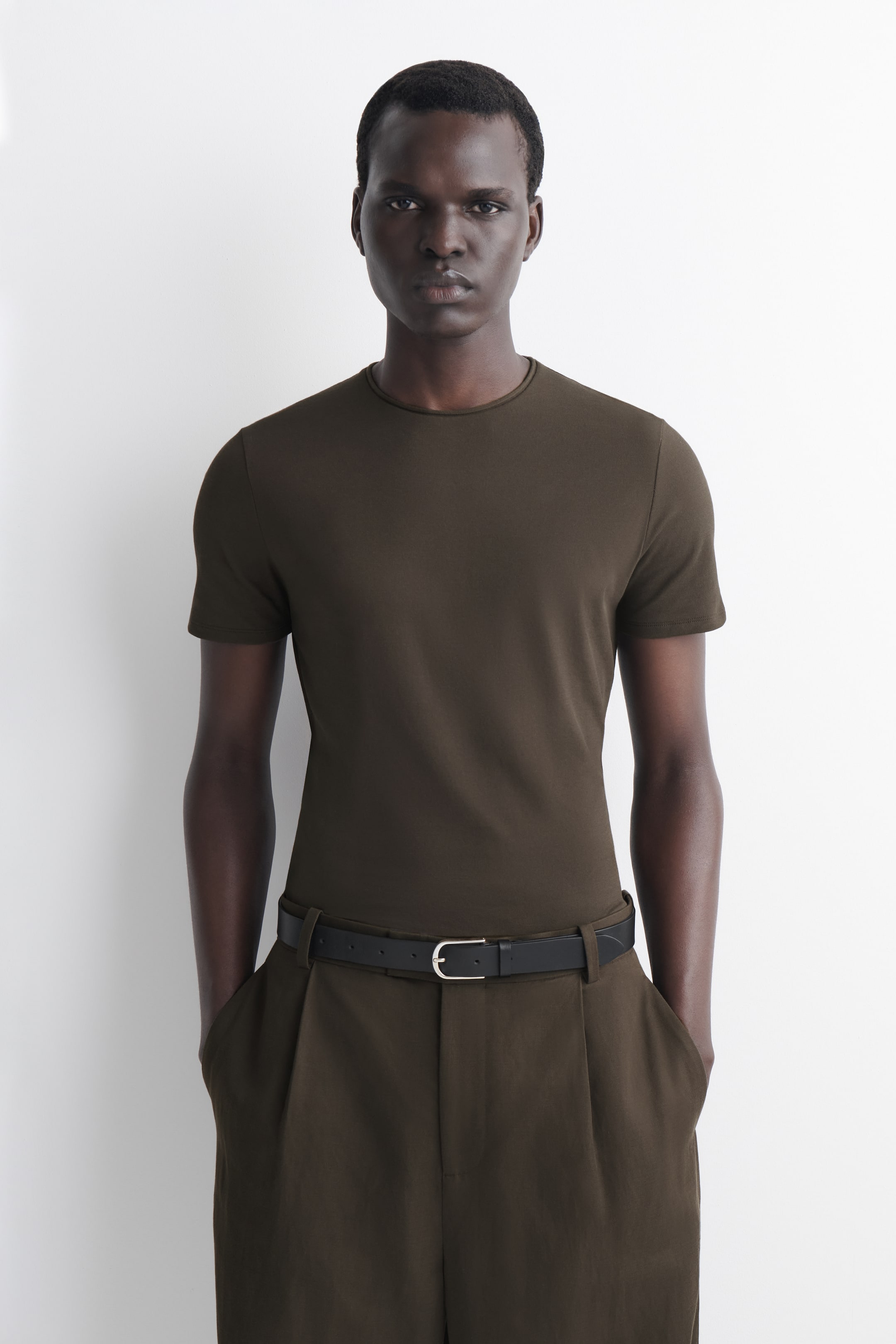 Agrandir l'image: Un t-shirt à col rond vert olive foncé à manches courtes est présenté, s'ajustant parfaitement au torse. Il est assorti à un pantalon plissé vert olive foncé, maintenu par une ceinture noire avec une boucle argentée.