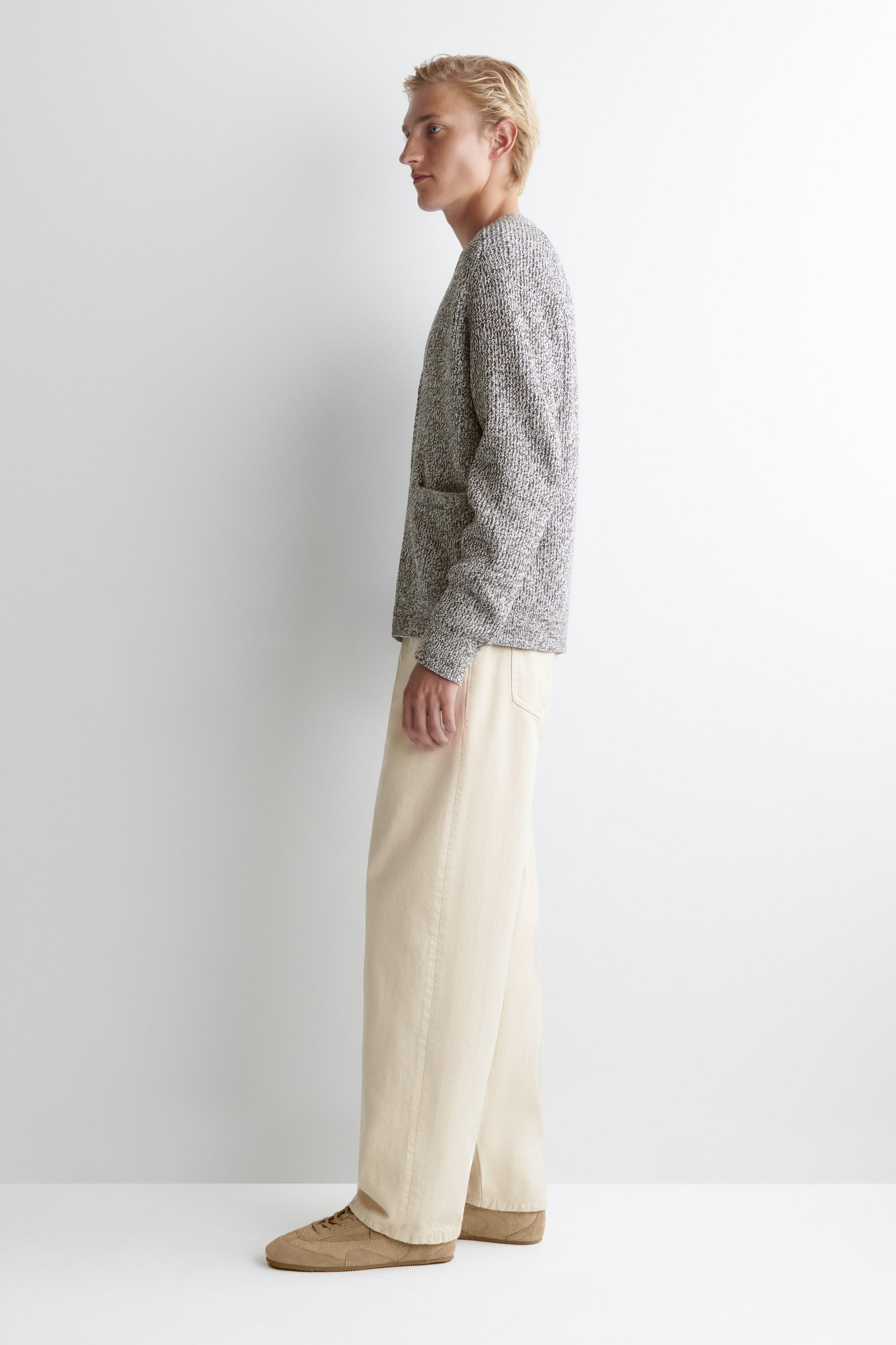Agrandir l'image: Un cardigan en maille gris chiné à coupe décontractée, avec une ouverture à l'avant et une poche plaquée visible, est présenté de profil, porté sur un pantalon beige clair à jambes larges tombant librement sur des baskets en daim couleur fauve.