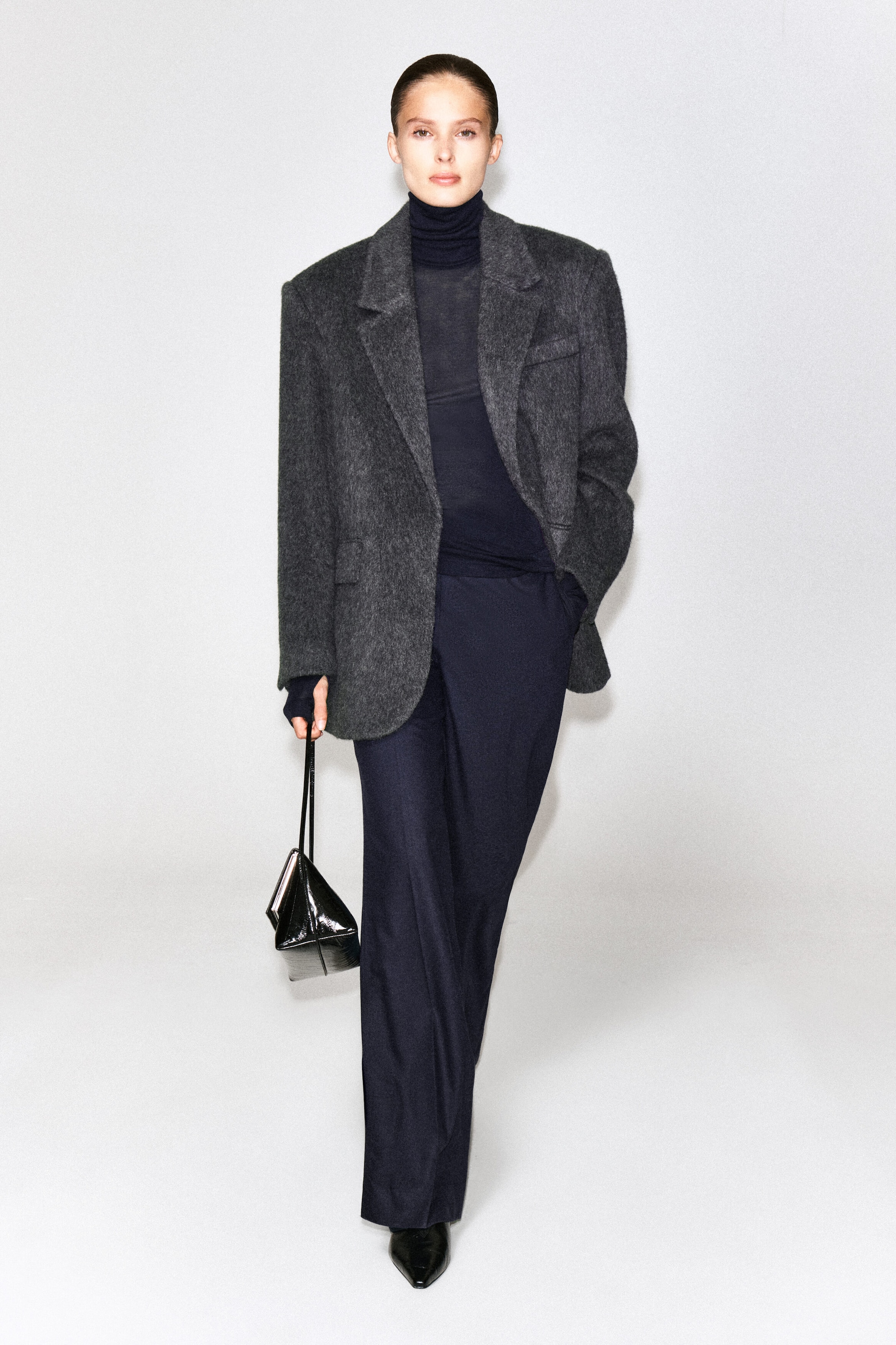 The navy merino wool high-neck top is worn by the model, styled as a layer underneath a textured dark gray blazer and tucked into dark trousers. The top features a fine-knit texture and a fitted turtleneck collar.