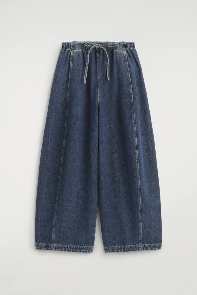Donkergewassen denim broek wordt gepresenteerd in een VOORAANZICHT, met een ontspannen pasvorm en volumineuze tonvormige pijpen. Ze zijn ontworpen met een elastische tailleband, tonale koorden die aan de voorkant zijn vastgebonden, schuine zijzakken en subtiele off-centre naden die langs de benen lopen. - 1
