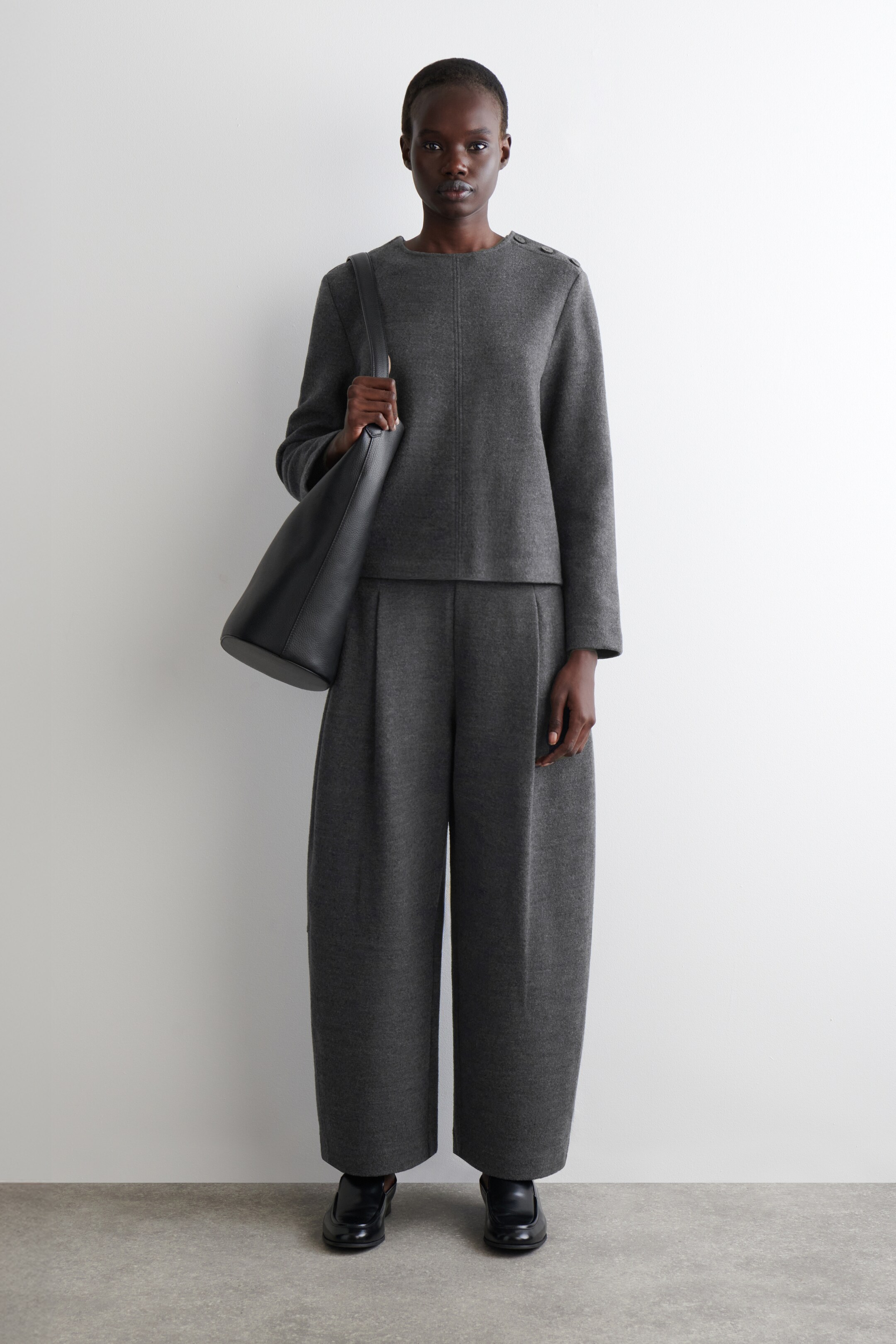 Agrandir l'image: Une tenue deux pièces grise est présentée, comprenant un haut à col rond orienté vers l'avant avec trois boutons sur chaque épaule et une couture verticale sur le devant, assorti à un pantalon large plissé. Un sac seau texturé noir est porté sur l'épaule droite, et des chaussures noires à enfiler sont portées.