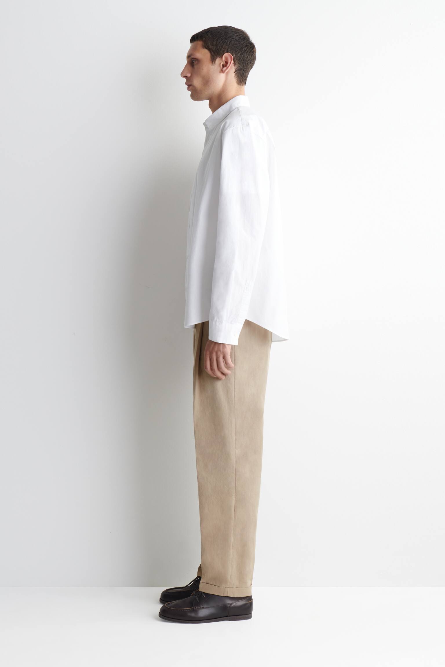 Une tenue est présentée de profil, mettant en avant une chemise blanche à manches longues et à boutons, avec une coupe décontractée. La chemise est associée à un pantalon plissé kaki, retroussé au-dessus de chaussures mocassins à lacets de couleur marron foncé. - 1