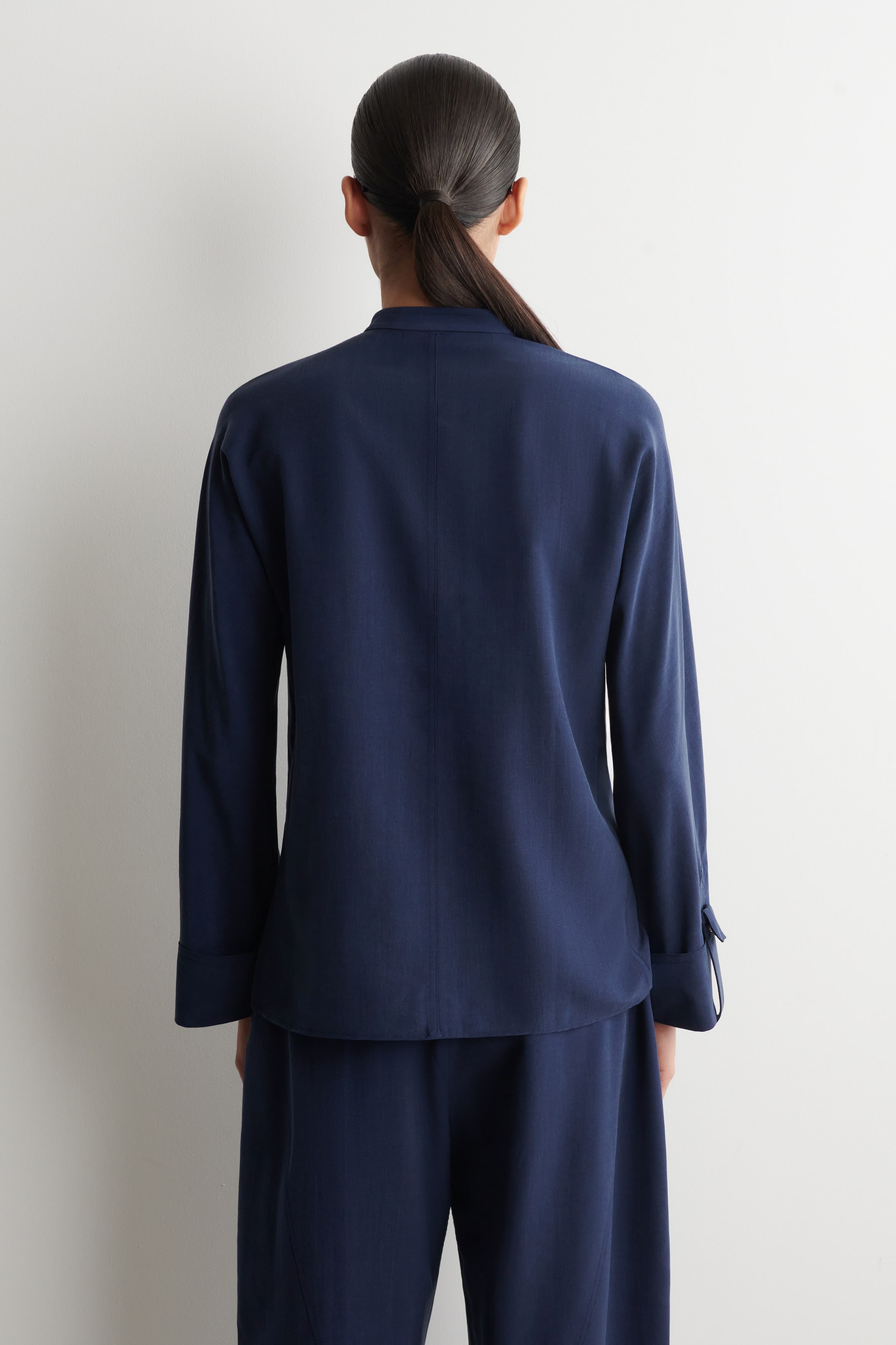 Ver imagen más grande: Una camisa azul oscuro fluida se muestra desde atrás, con un cuello de banda y mangas largas con lengüetas que aseguran los puños.